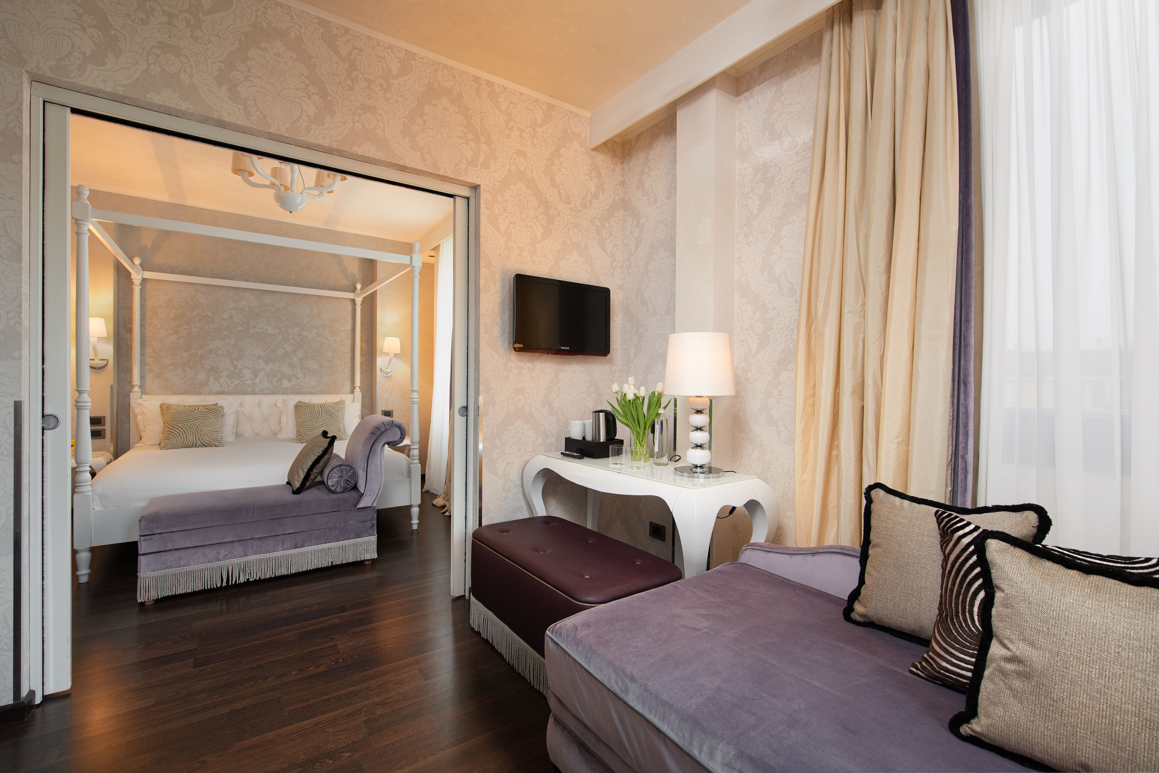 A hotel room with a bed, a sofa, a small table with a lamp, and a TV on the wall.