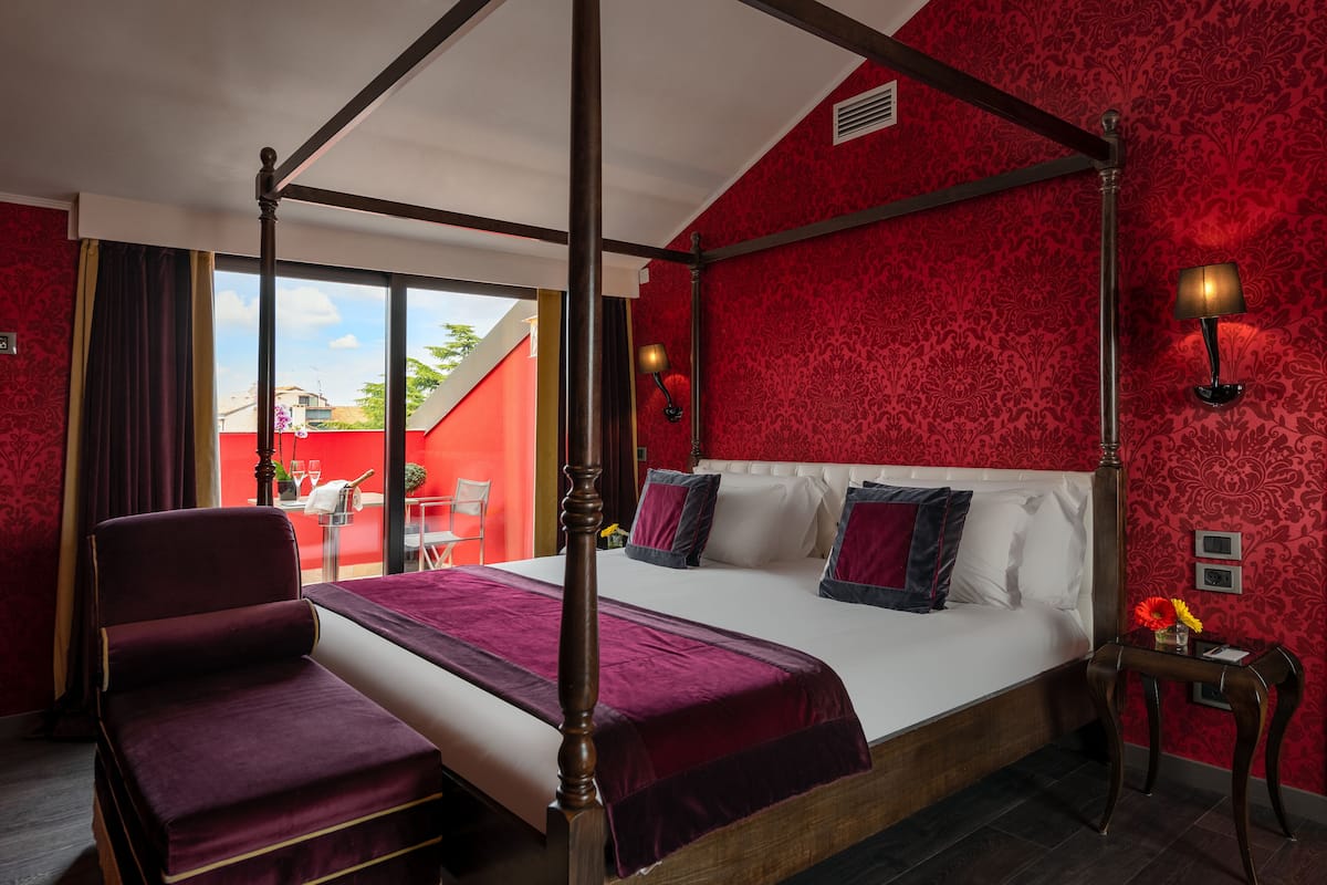 A four-poster bed with a red and white color scheme, a wooden chair, and a small table with a lamp.