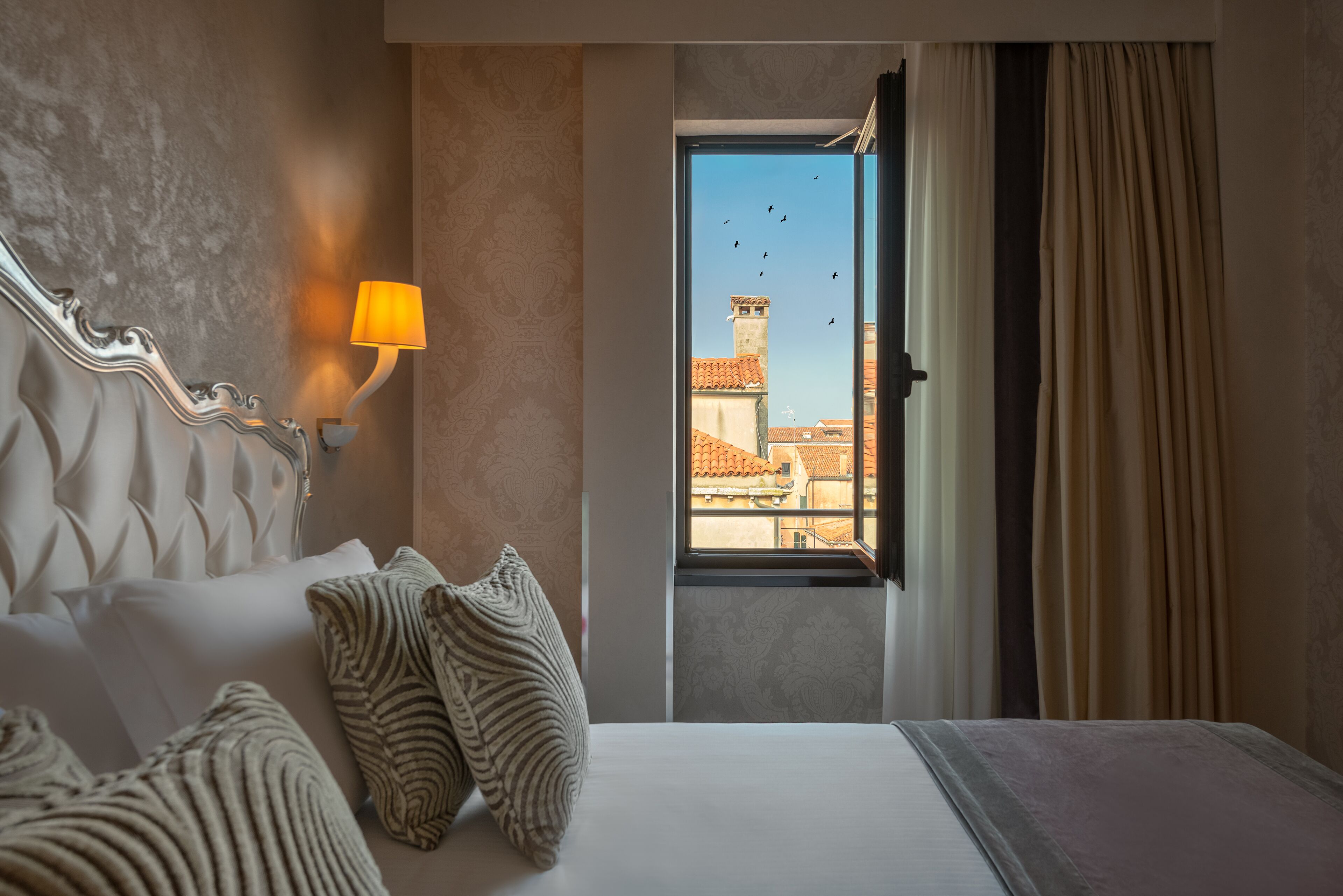 Habitación de hotel con cama, almohadas, una lámpara de pared y una ventana que ofrece vistas a edificios y aves.