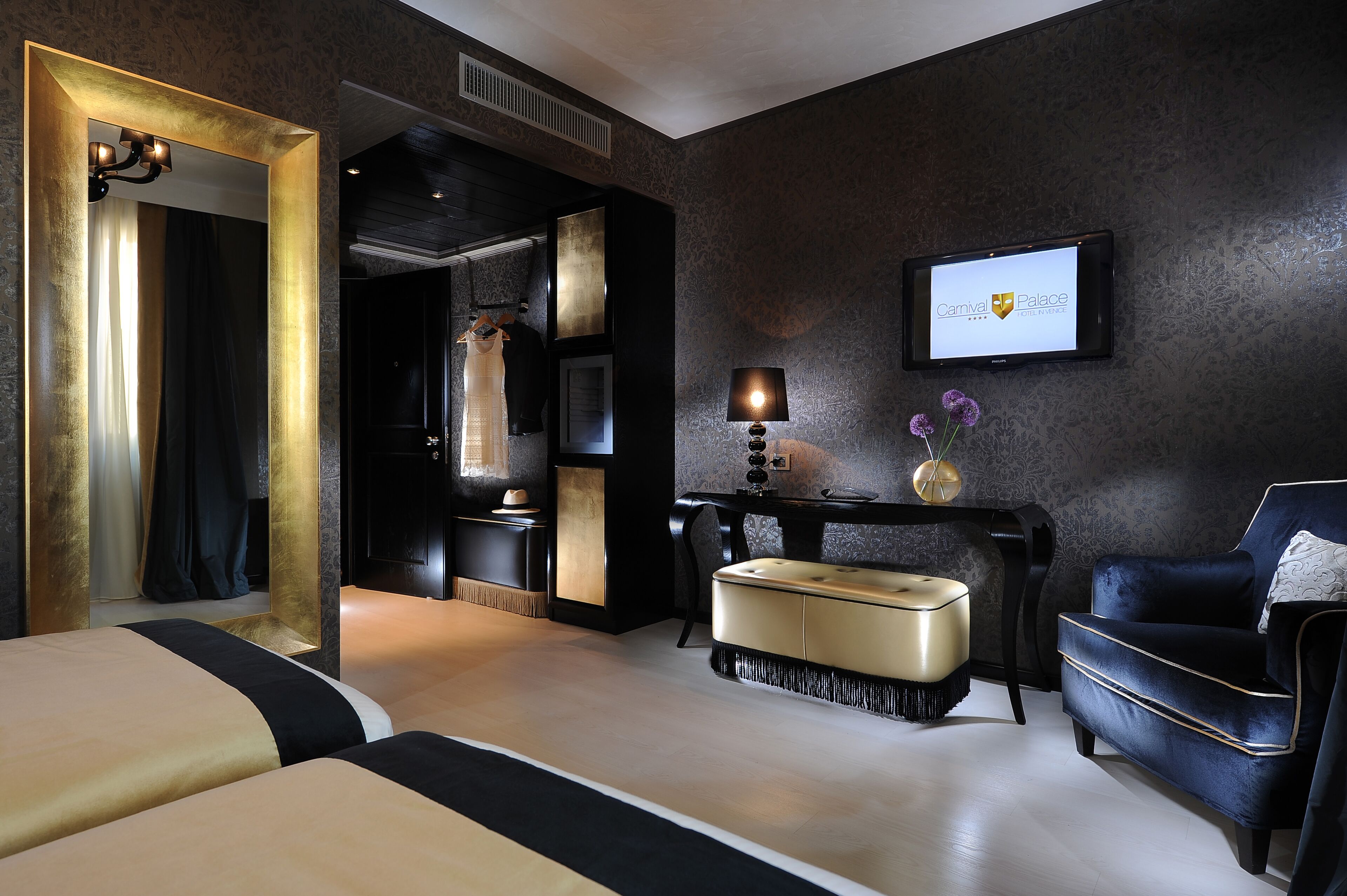 Una habitación de hotel moderna con un espejo grande, una televisión mostrando el nombre del hotel, un sofá y una mesita de noche con una lámpara.