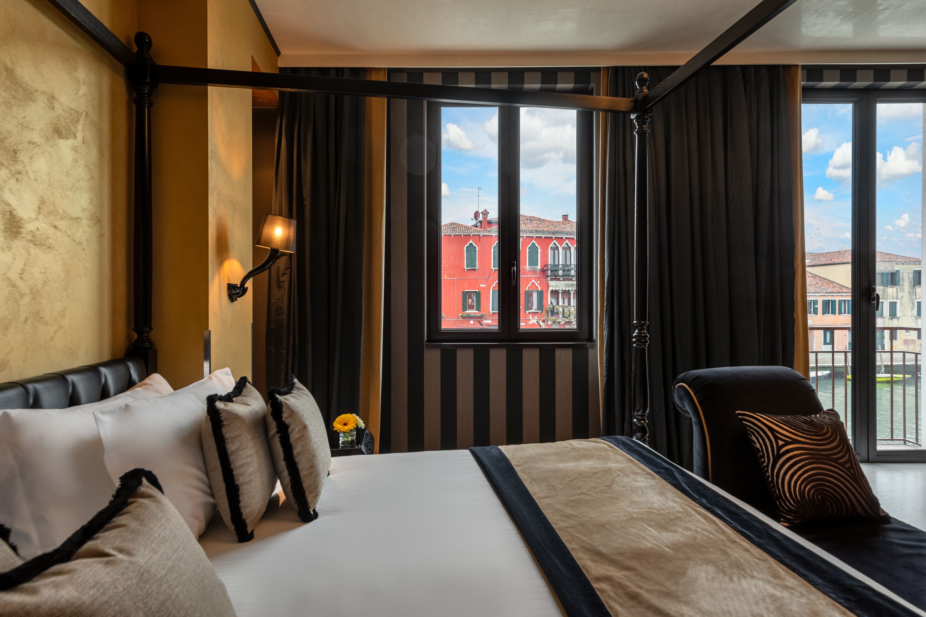A hotel room with a large bed, black and white striped curtains, a view of a red building, and a black armchair.