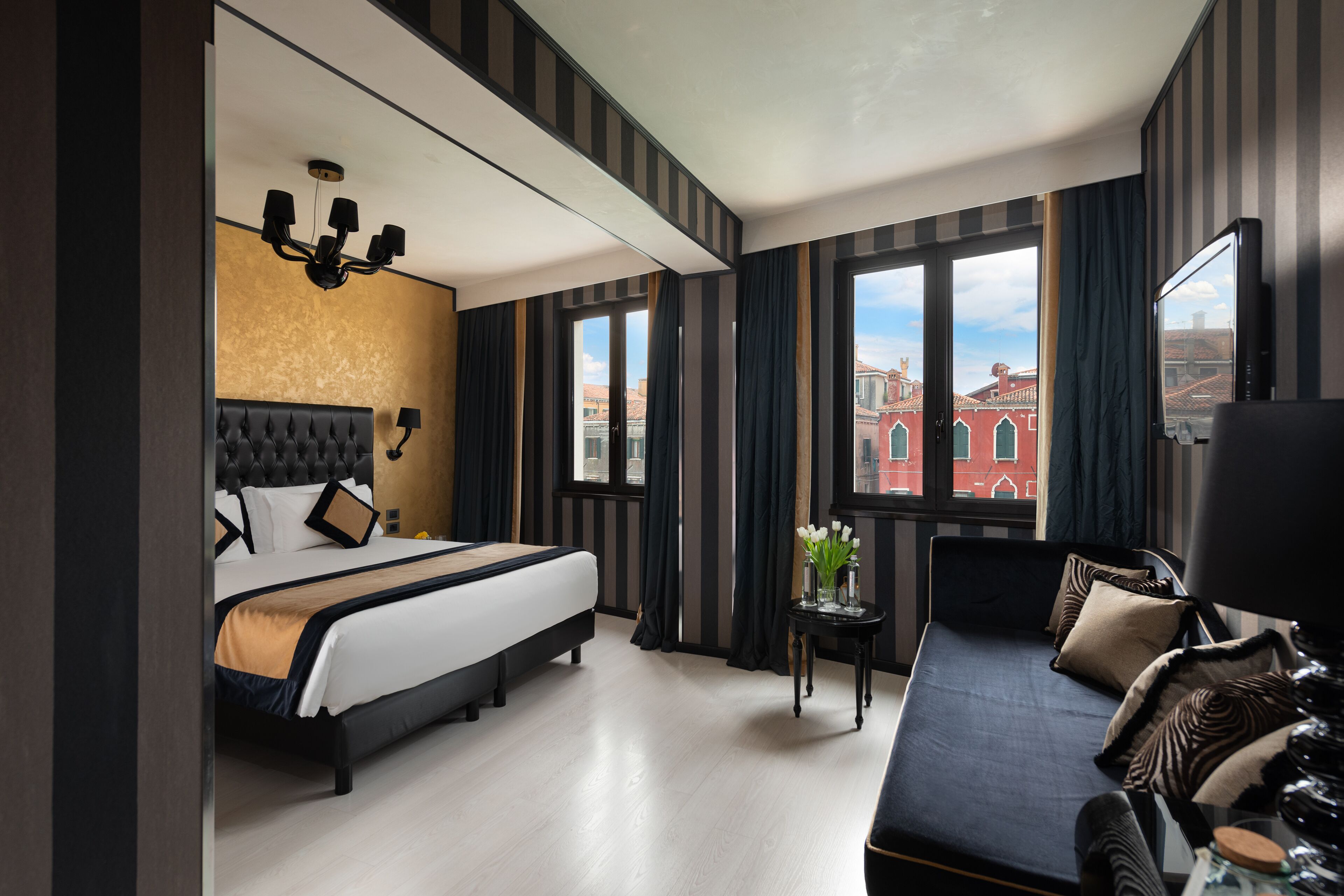 A hotel room with a large bed, a sofa, a small table with a vase of flowers, and a view of a red building through the windows.