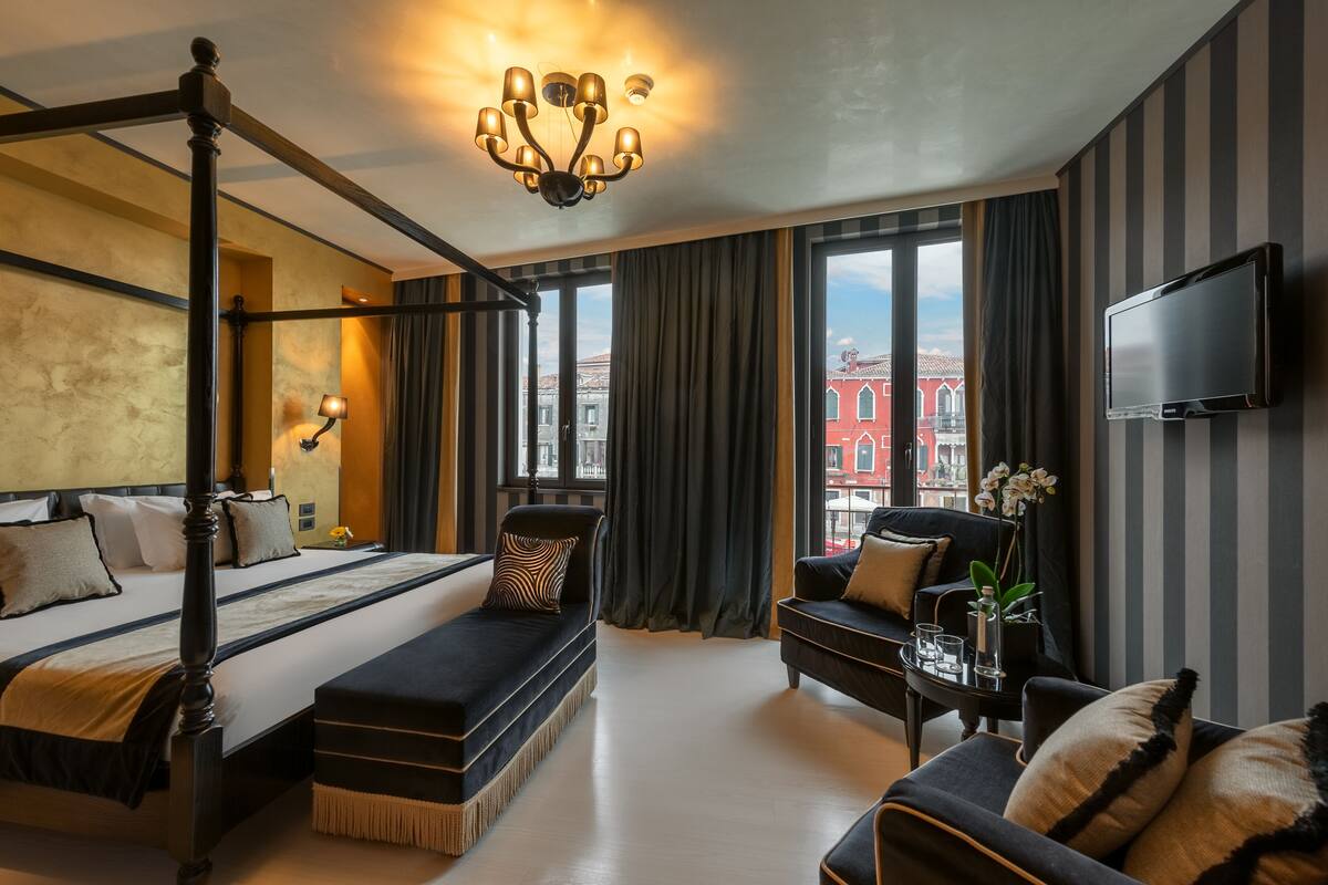 A four-poster bed with a bench, a sofa, a TV, and a view of a red building through large windows.