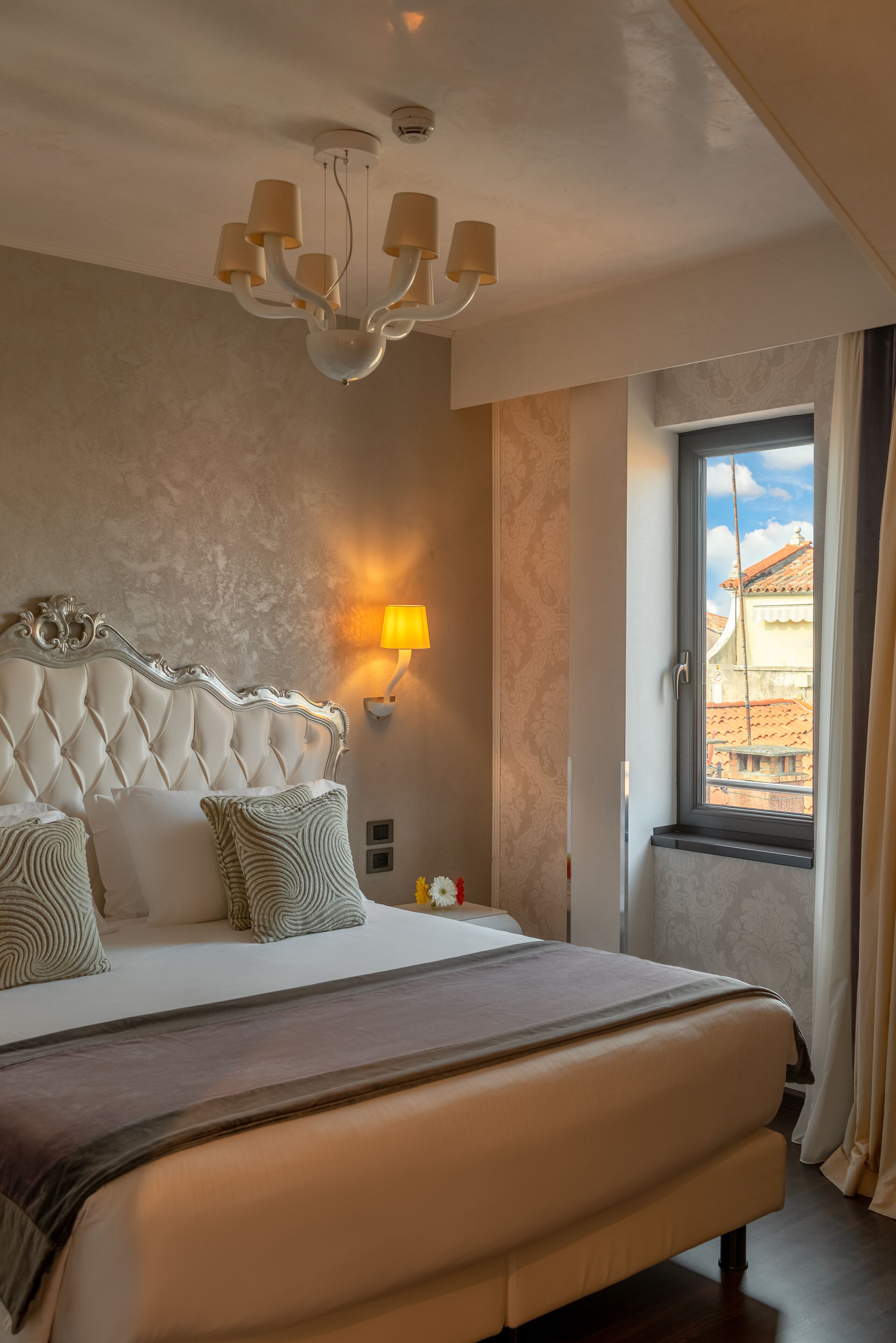 A bedroom with a tufted headboard, a large bed, a nightstand with a lamp, and a window with a view of buildings.