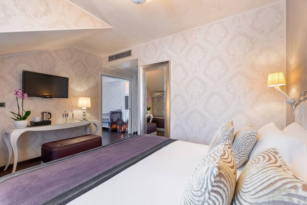 A modern hotel room with a large bed, a desk with a flat-screen TV, a mirror, and a bathroom visible through an open door.