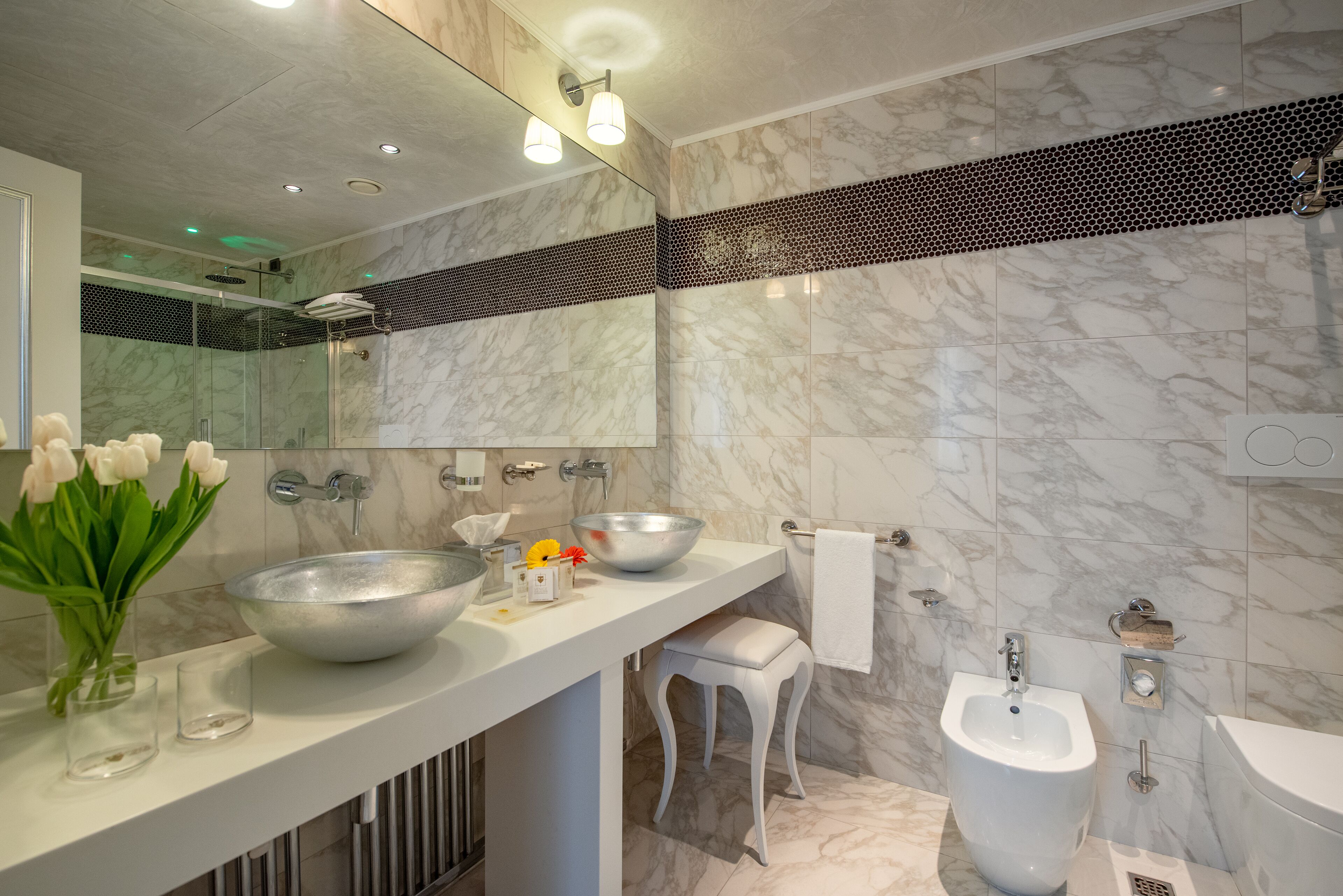 Un baño moderno con un espejo grande, un lavamanos blanco, un bidé y un revestimiento de mármol en la pared.