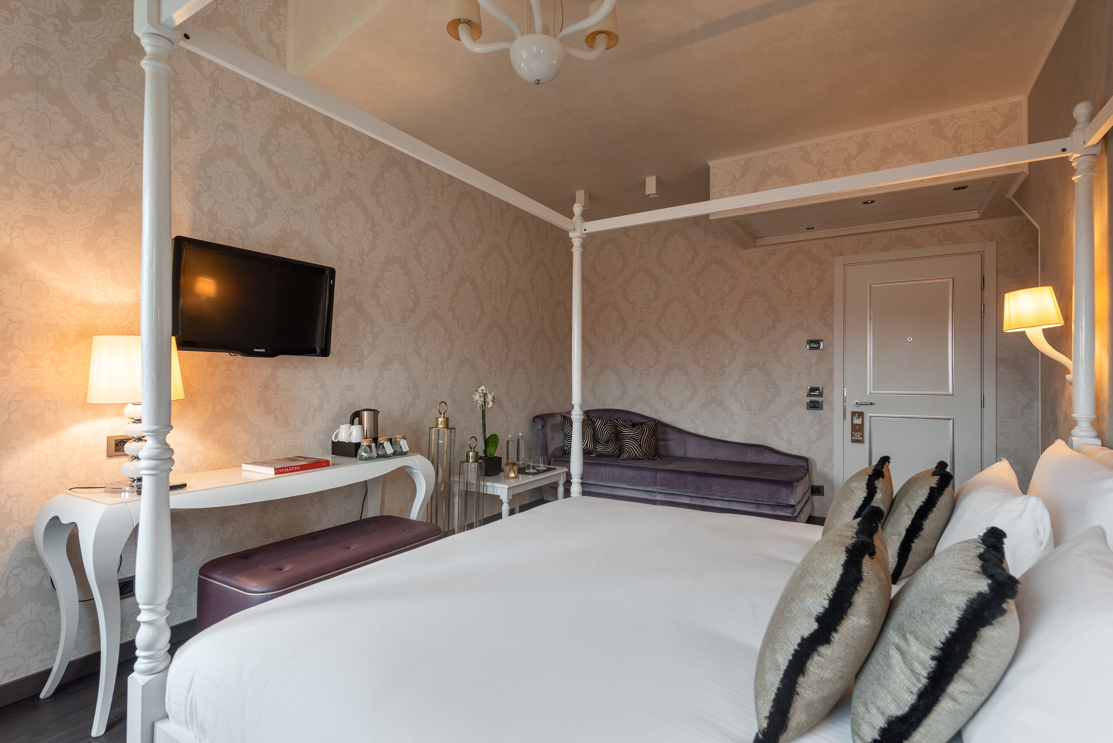 A four-poster bed with a flat-screen TV mounted above it, a bedside table with a lamp, a small table with a vase and a book, and a purple sofa against the wall.