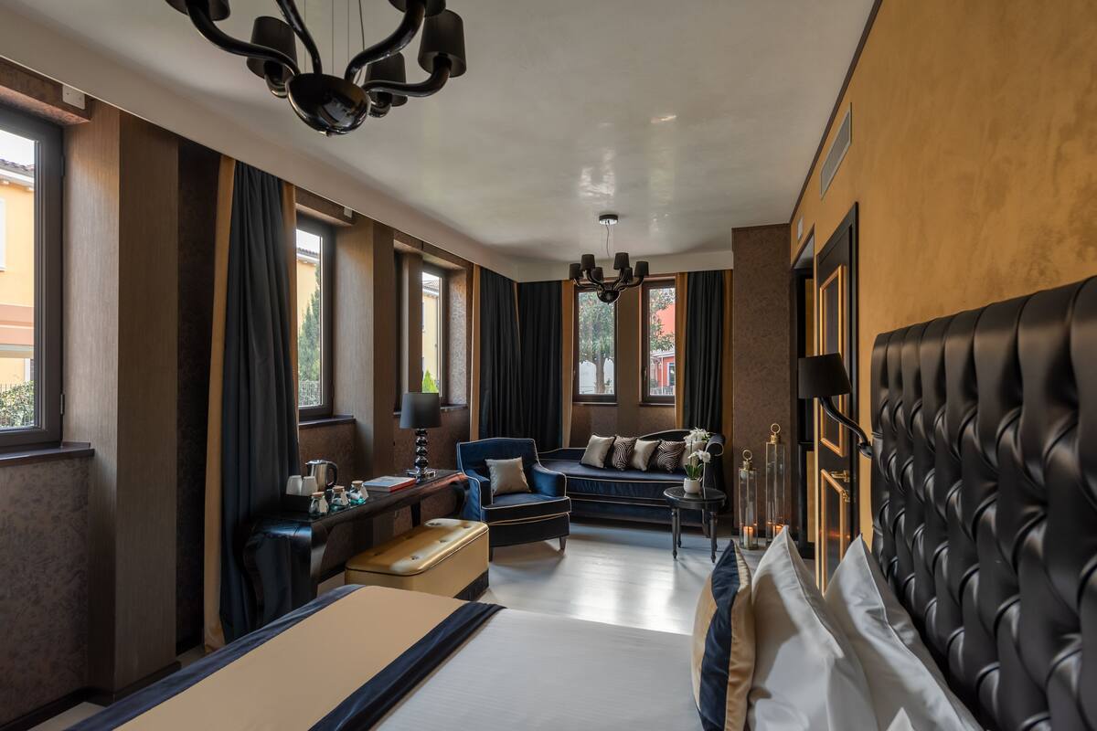 A hotel room with a bed, a sofa, a desk with a lamp, and a chandelier.
