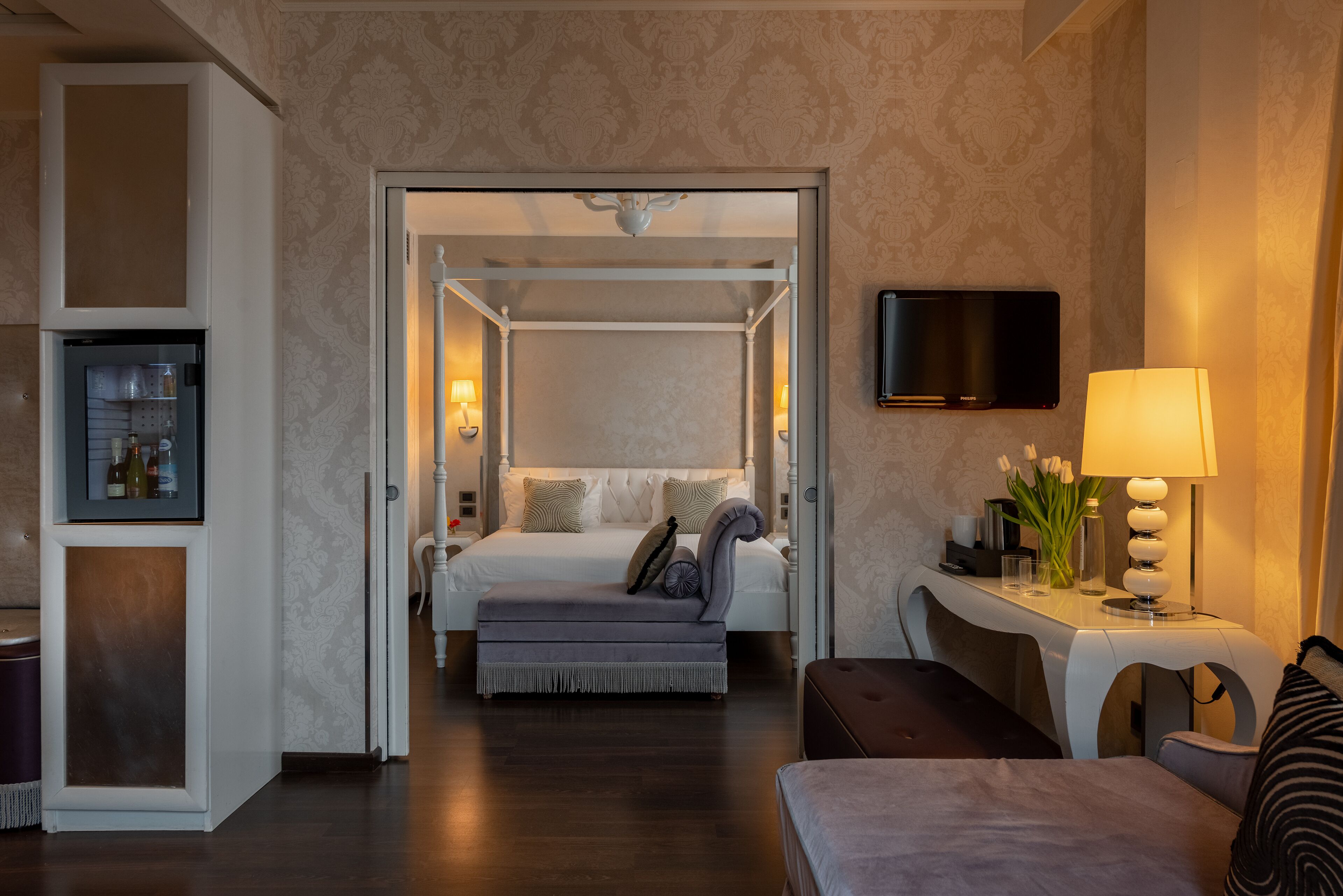 A hotel room with a bed, a desk with a lamp, a television, and a minibar.