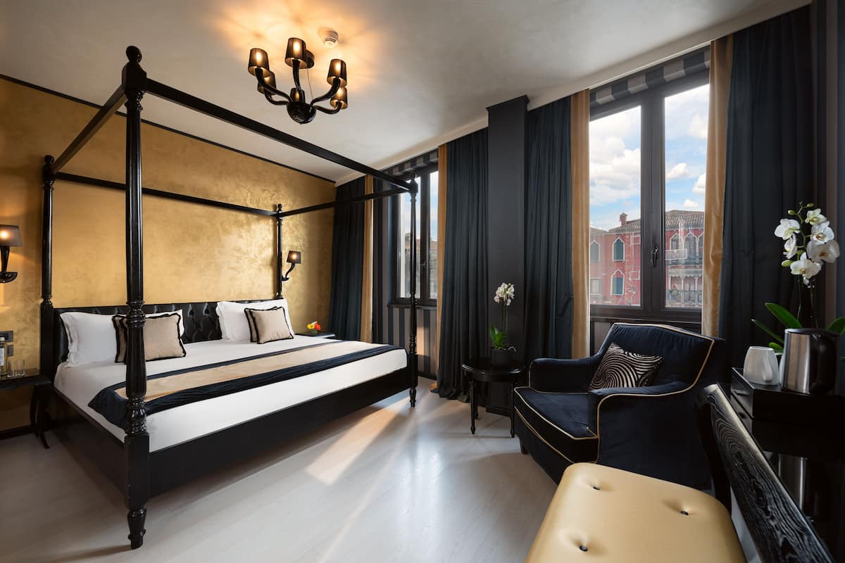 A four-poster bed with a black frame and white bedding, a dark blue armchair, a small side table, and a view of a red building through large windows.