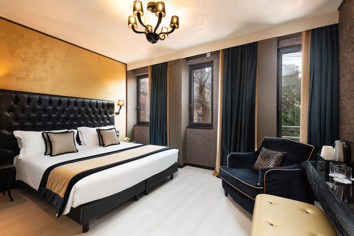 A modern hotel room with a large bed, a black upholstered armchair, a small sofa, and a dining table with a vase and glasses.