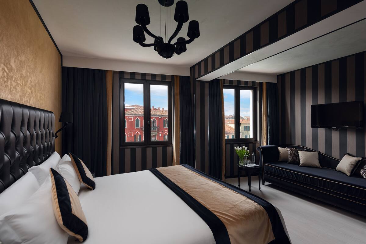 A hotel room with a large bed, a sofa, a TV, and a view of buildings outside the window.