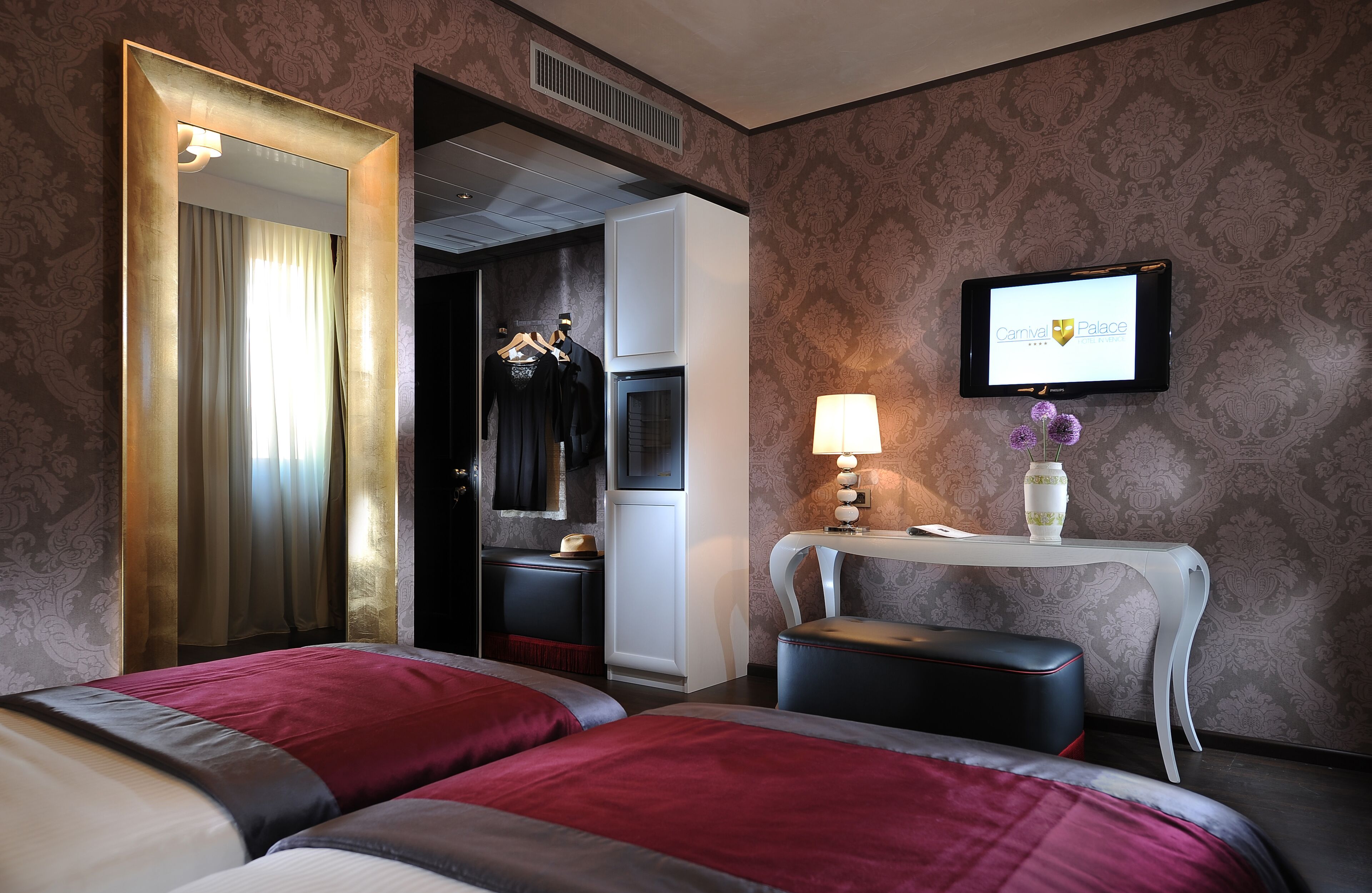 A hotel room with two beds, a vanity with a mirror, a television, and a small table with a lamp and decorative items.
