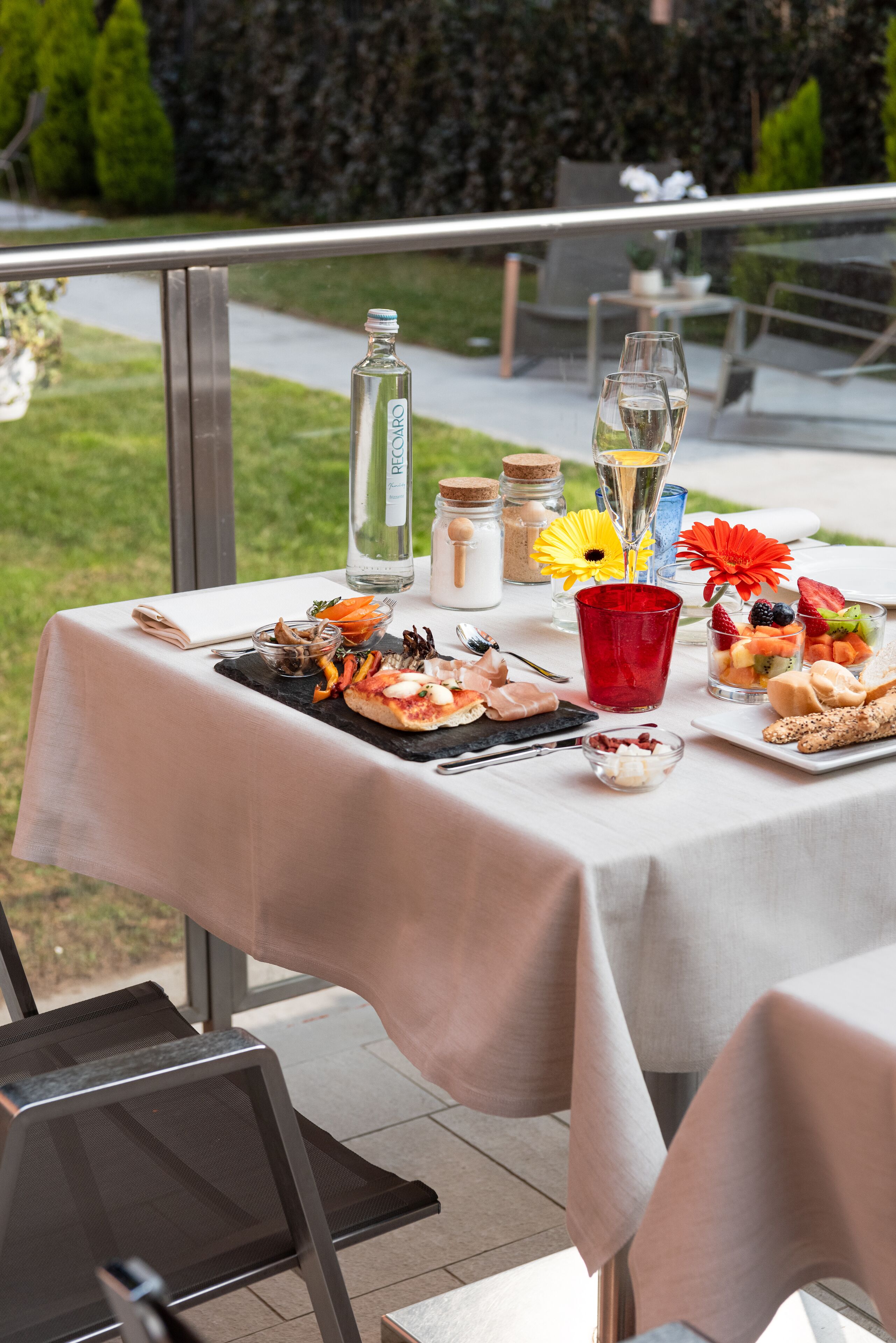 A table set outdoors with a variety of food and drinks, including a bottle of water, glasses, and a fruit platter.