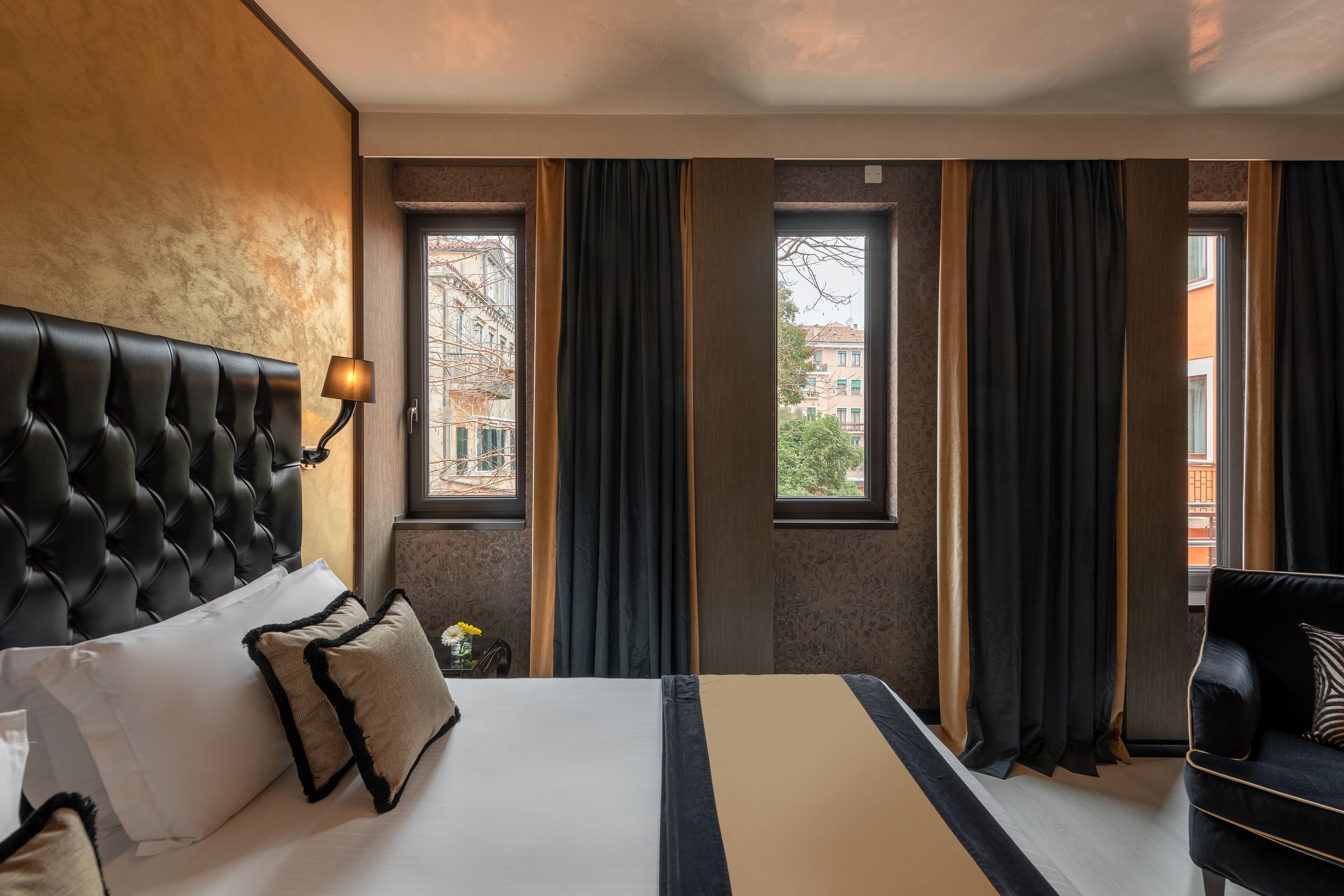 A hotel room with a tufted headboard, a large bed with white and beige bedding, a black armchair, and a view of buildings through windows with dark curtains.