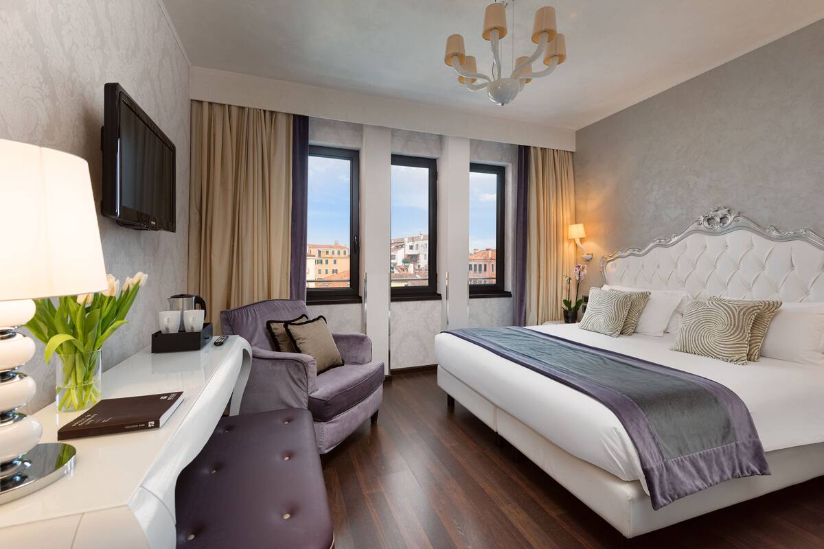 A modern hotel room with a large bed, a sofa, a desk with a lamp, and a TV mounted on the wall.