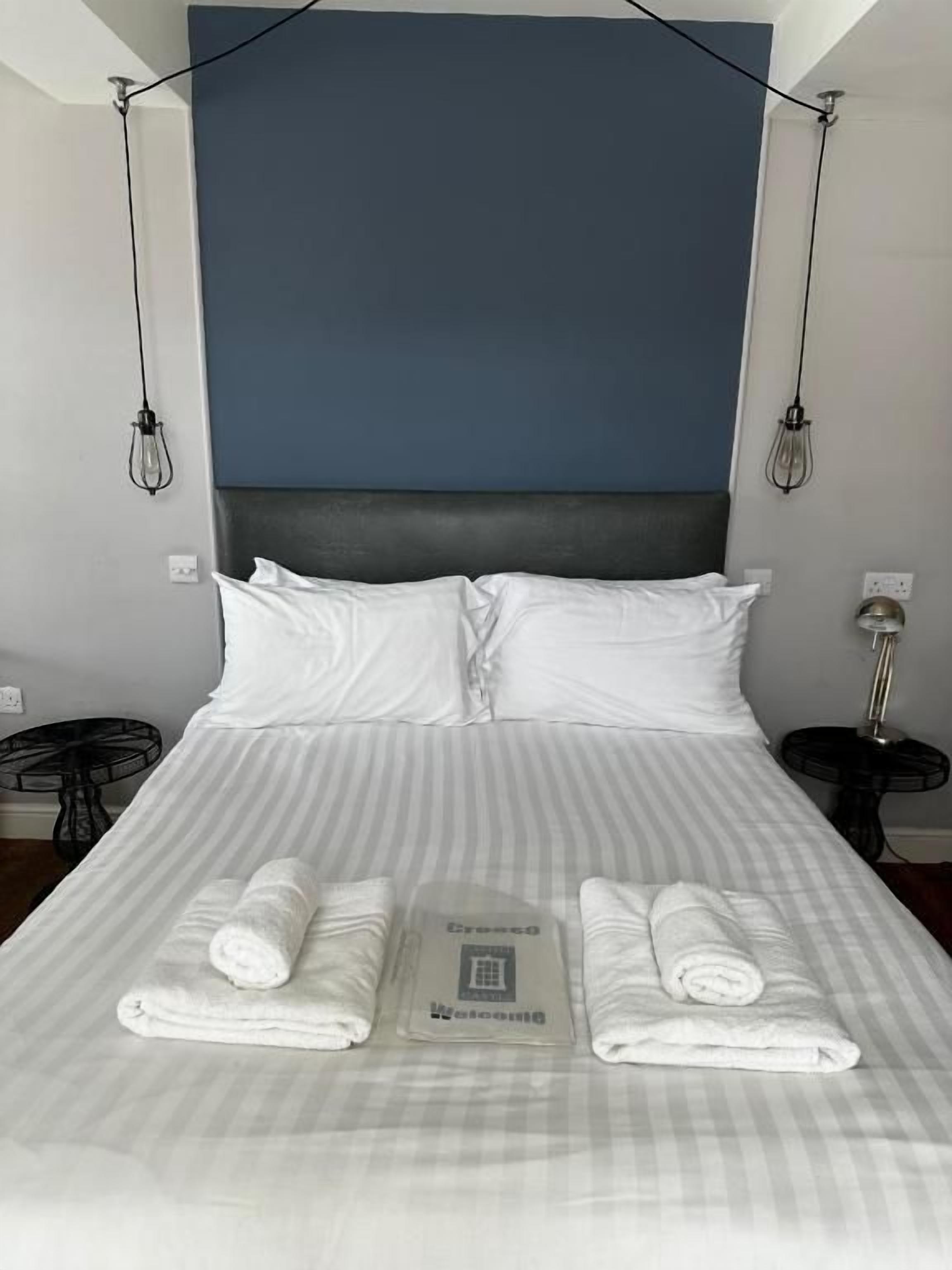 A neatly made bed with two rolled towels and a small booklet, a dark headboard, and a wall-mounted light fixture.