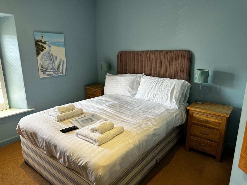 A neatly made bed with white linens and two towels, a bedside table with a lamp, and a painting of a beach on the wall.