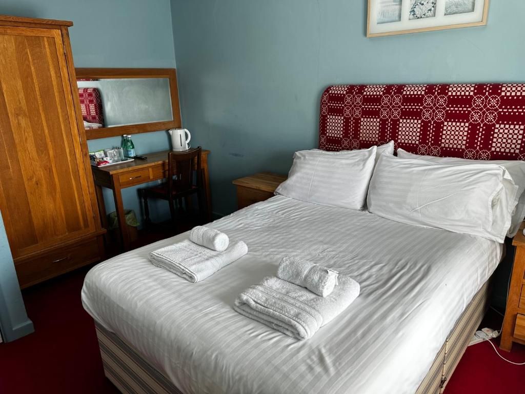 Una cama bien tendida con ropa blanca y toallas, un armario de madera, un escritorio con un hervidor y un cabecero estampado.