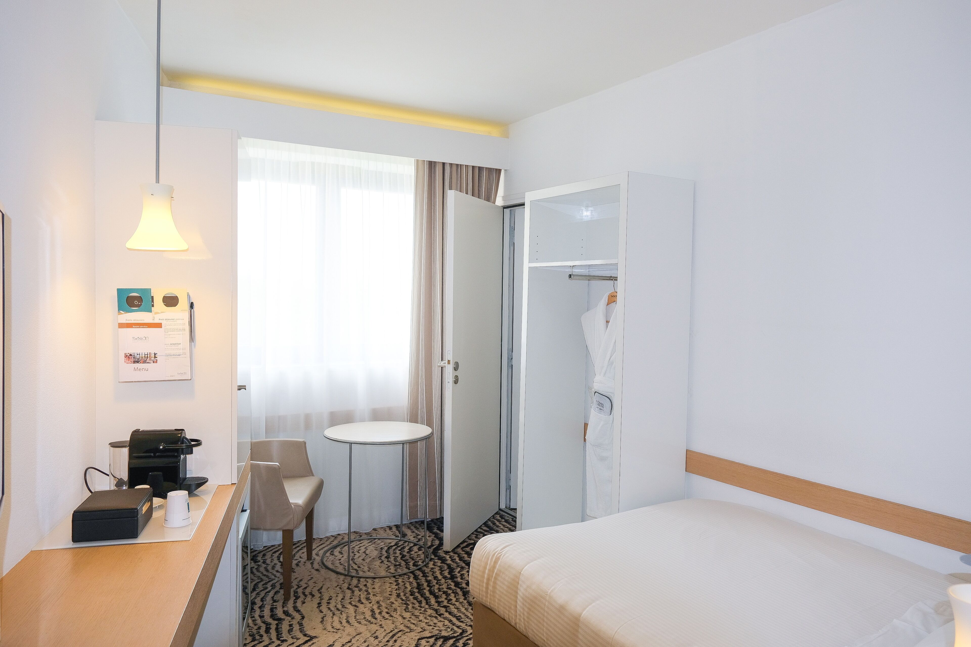 A hotel room with a bed, a desk with a coffee maker, a chair, a wardrobe, and a small table.
