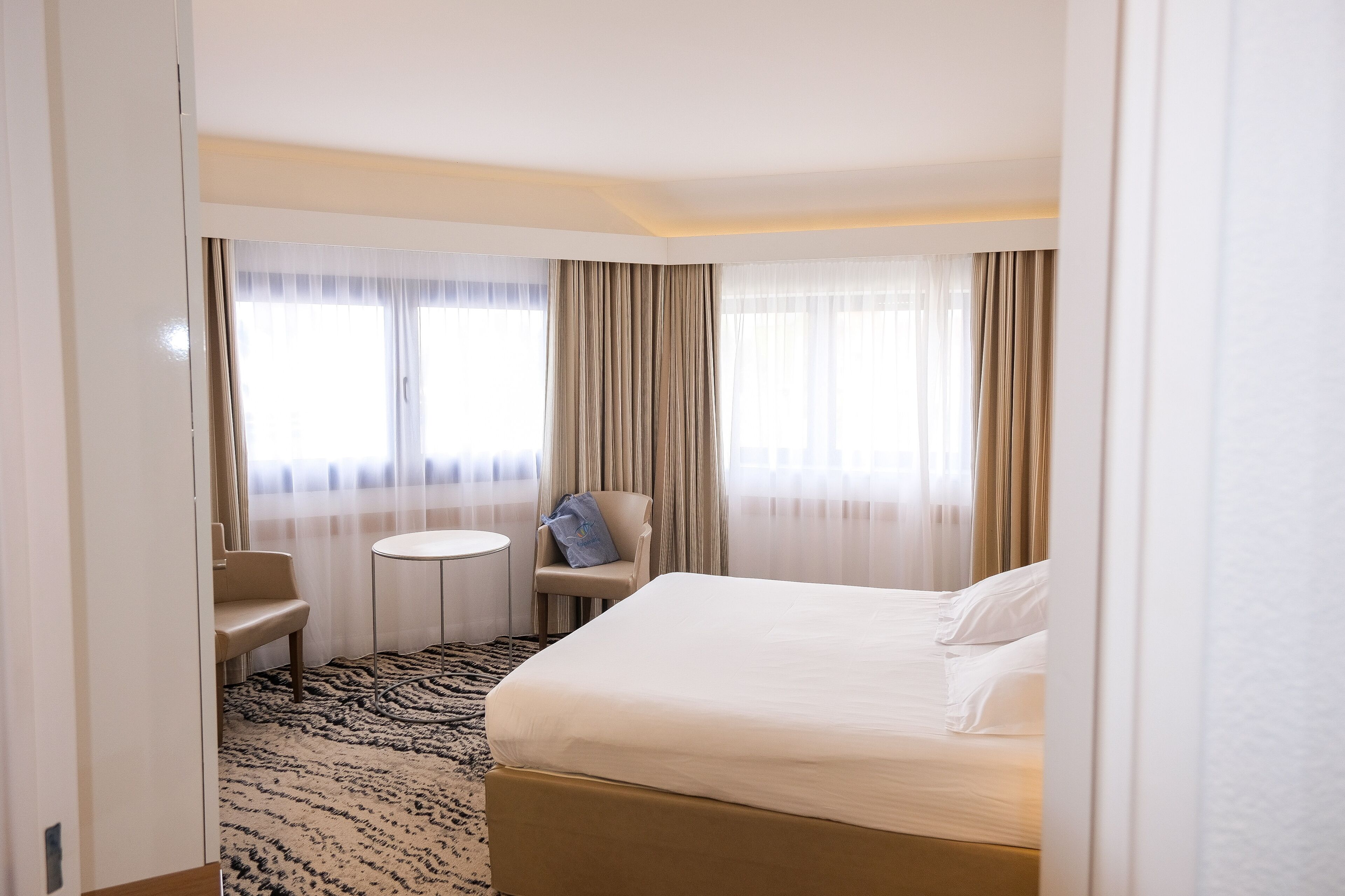 Una habitación de hotel con cama, una silla, una mesita y un ventanal con cortinas.