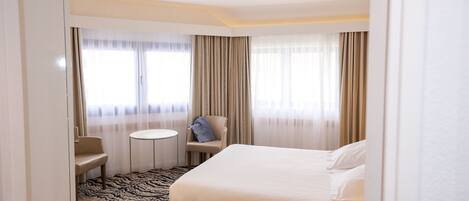 Una habitación de hotel con cama, una silla, una mesita y un ventanal con cortinas.
