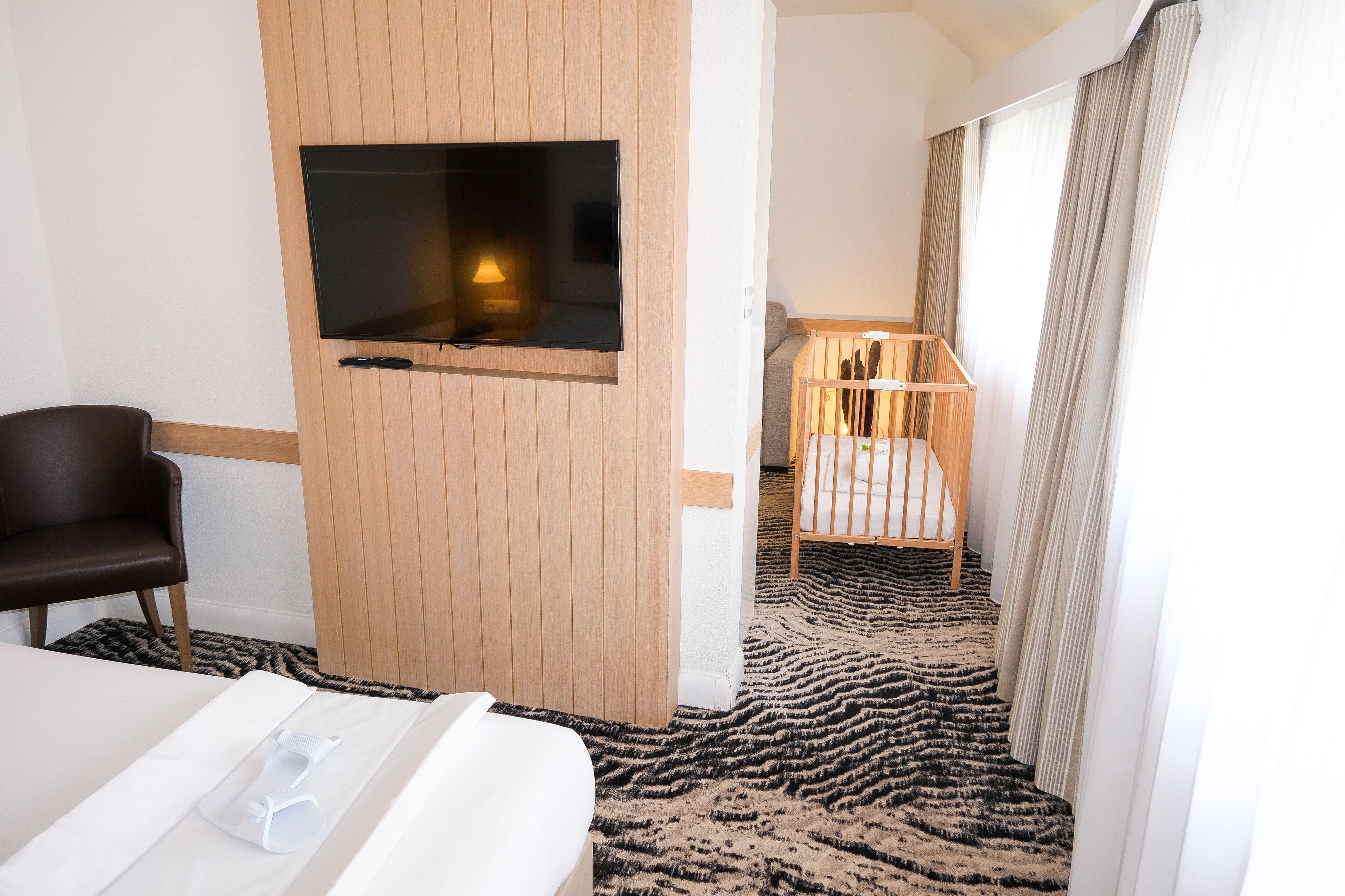 A hotel room with a bed, a TV mounted on the wall, a wooden crib, and a patterned carpet.