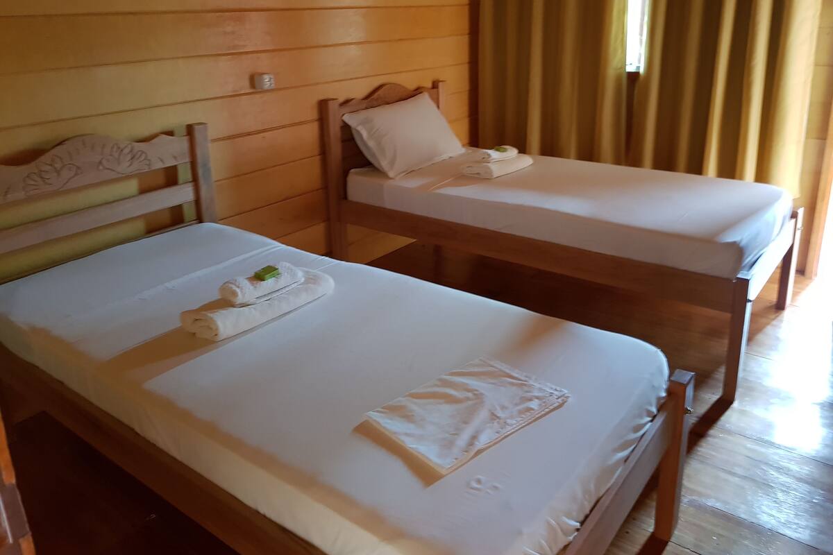Deux lits en bois avec du linge de lit et des serviettes blancs, une tête de lit en bois et une fenêtre avec des rideaux.