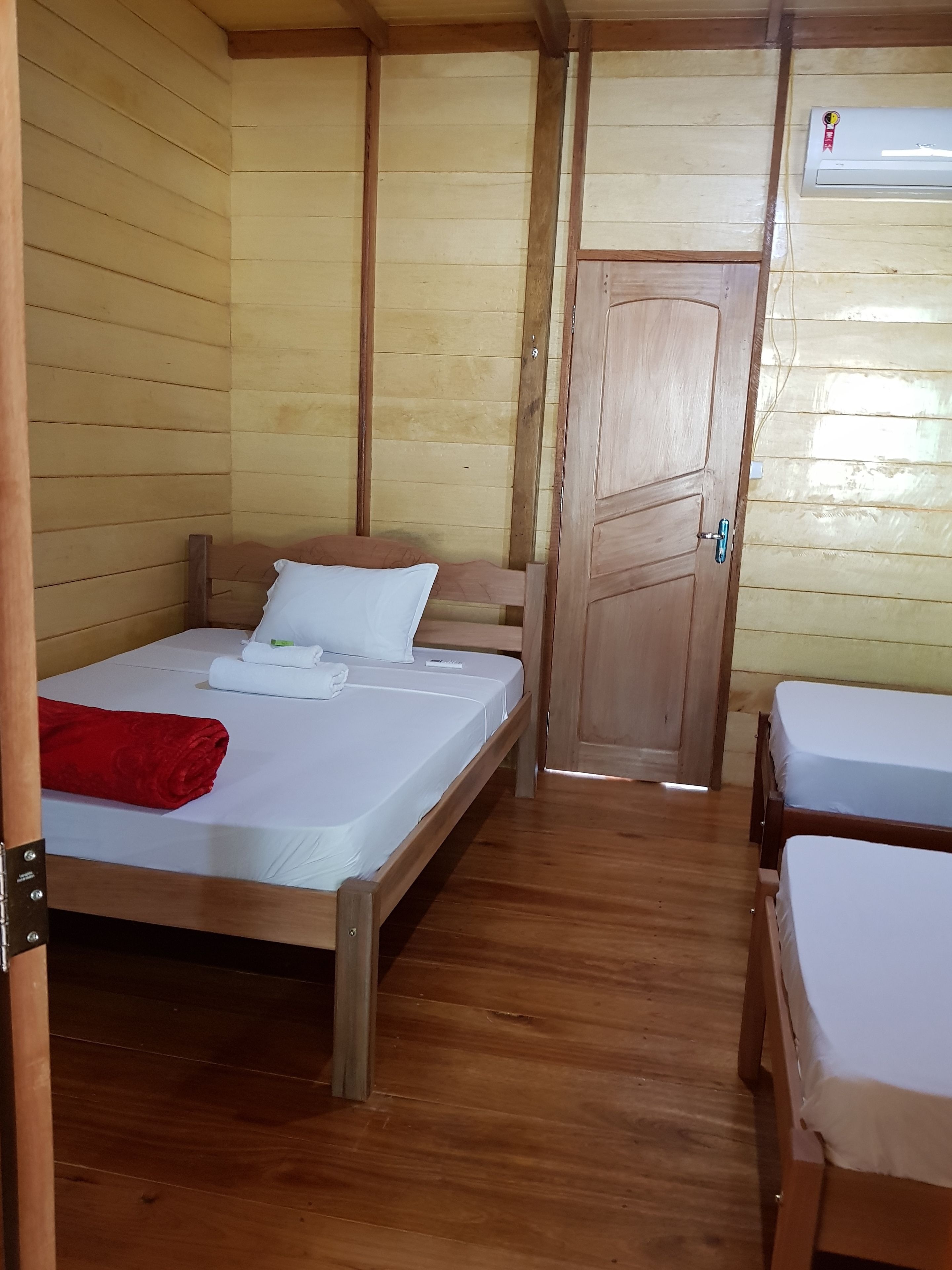 A room with wooden walls and floors, a bed with white bedding, a red towel, and a door leading to another room.