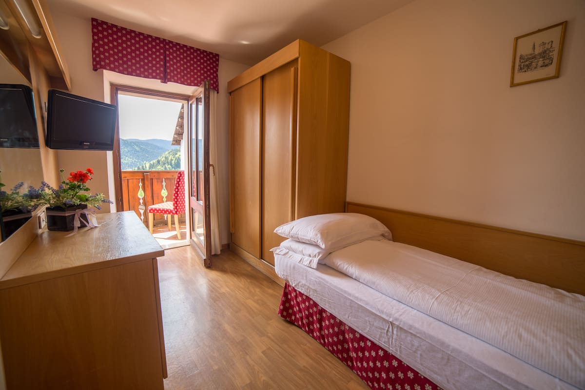 A bedroom with a bed, a wooden desk, a chair, a TV, and a balcony with a view of the mountains.