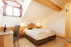 Ein Schlafzimmer mit einem Holzkopfende, zwei Betten, einem Schreibtisch mit Stuhl und einem kleinen Tisch mit Flasche und Obst.