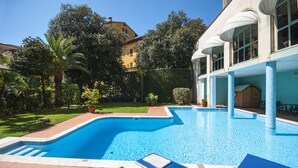 A swimming pool with blue water, surrounded by a garden with various plants and trees. There are lounge chairs and a wooden structure in the background. - Settentrionale Esplanade (Montecatini Terme)