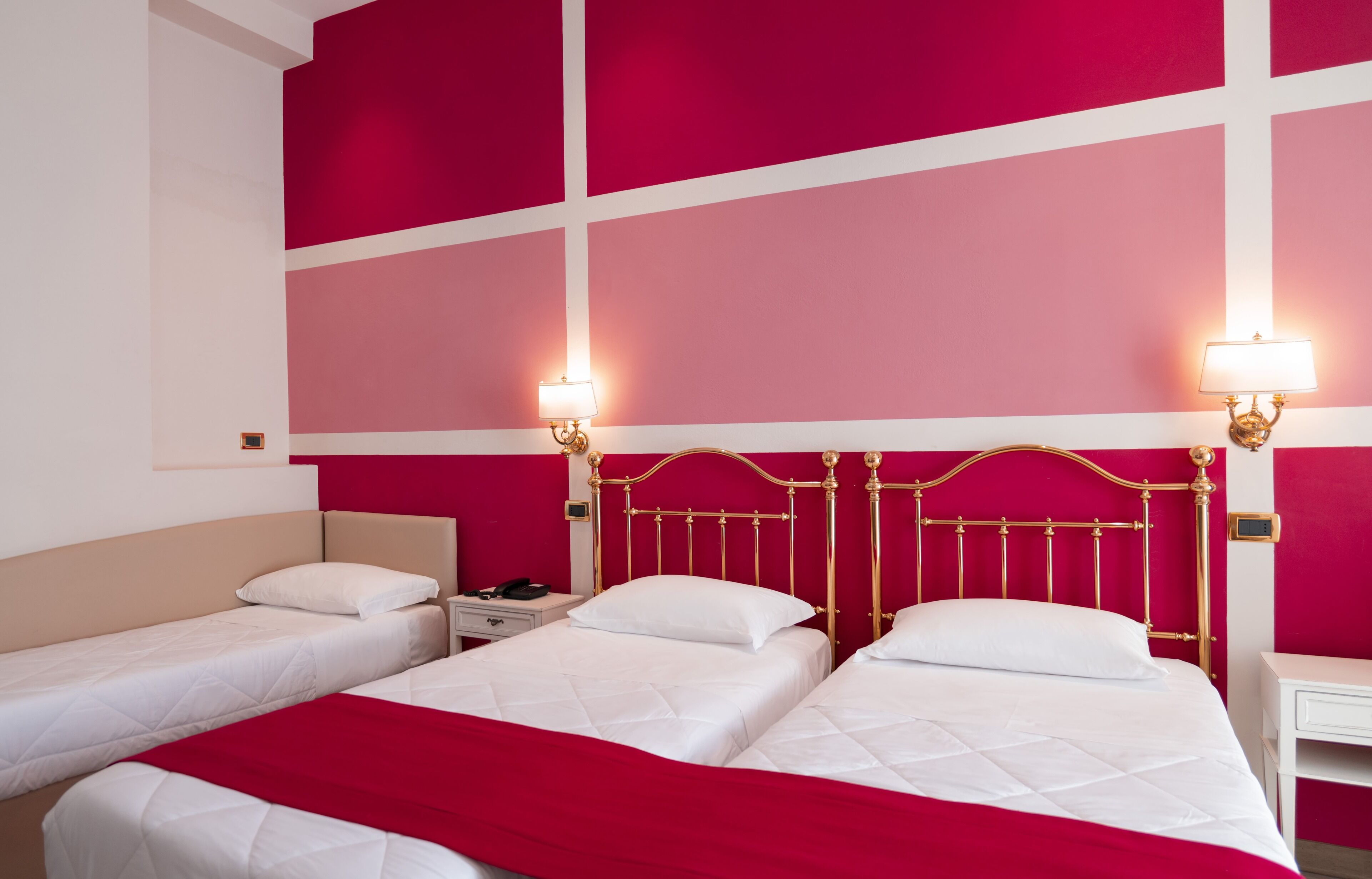 A hotel room with two beds, a red accent wall, and two wall-mounted lamps.