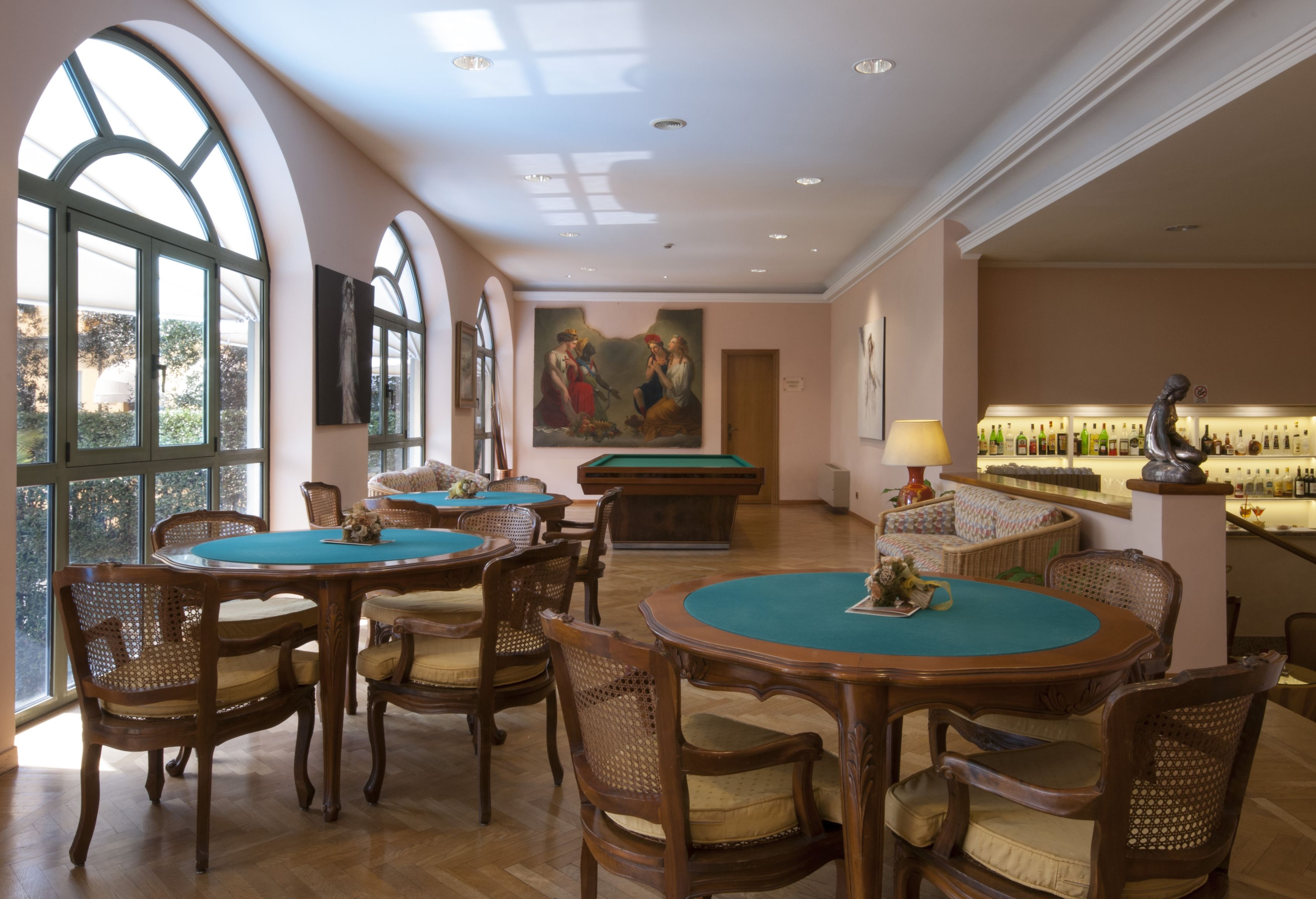 A billiards room with multiple tables and chairs, a pool table, a bar area with bottles, and a large painting on the wall.