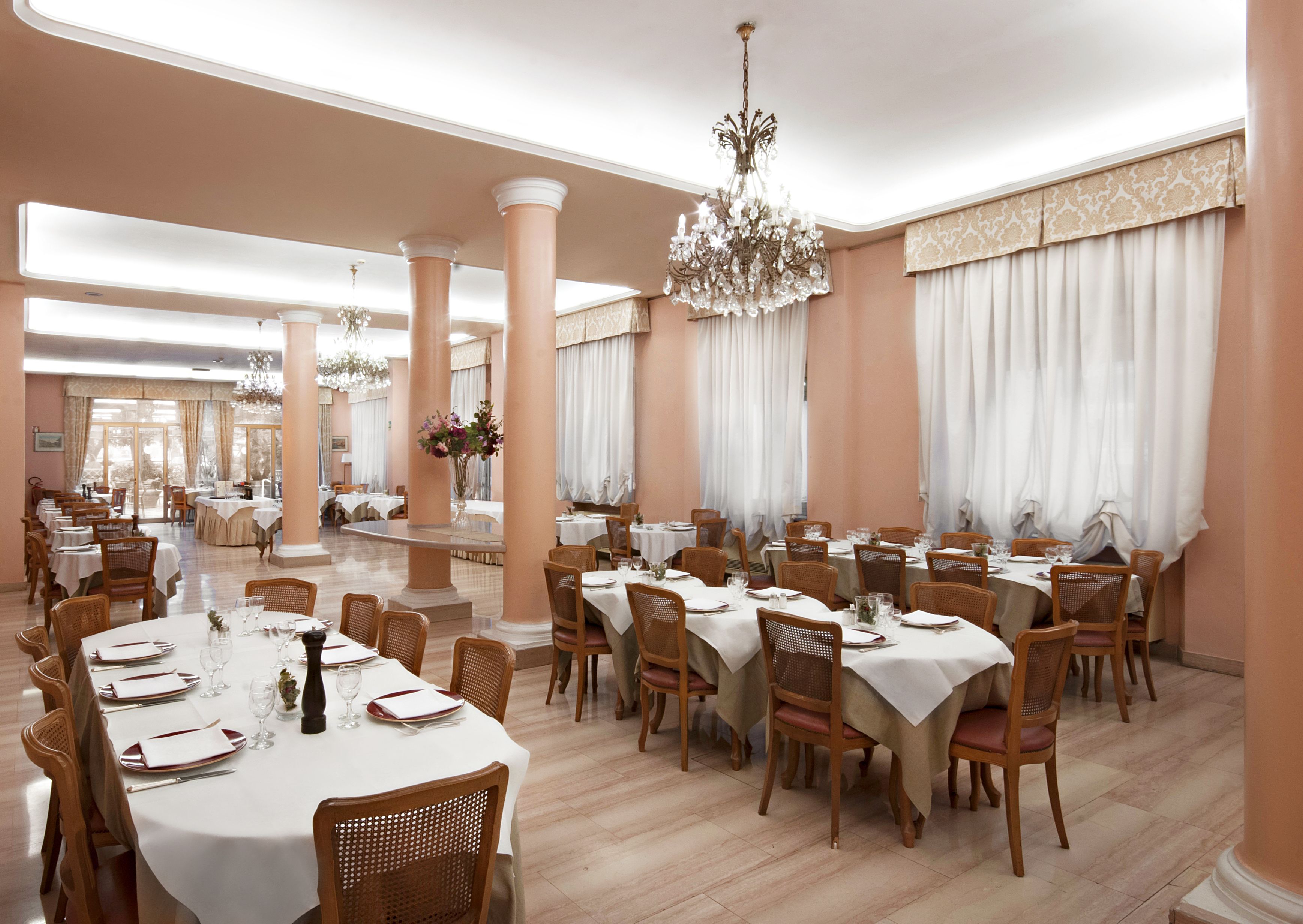 A spacious dining area with multiple tables set for service, elegant chandeliers, and large windows with curtains.