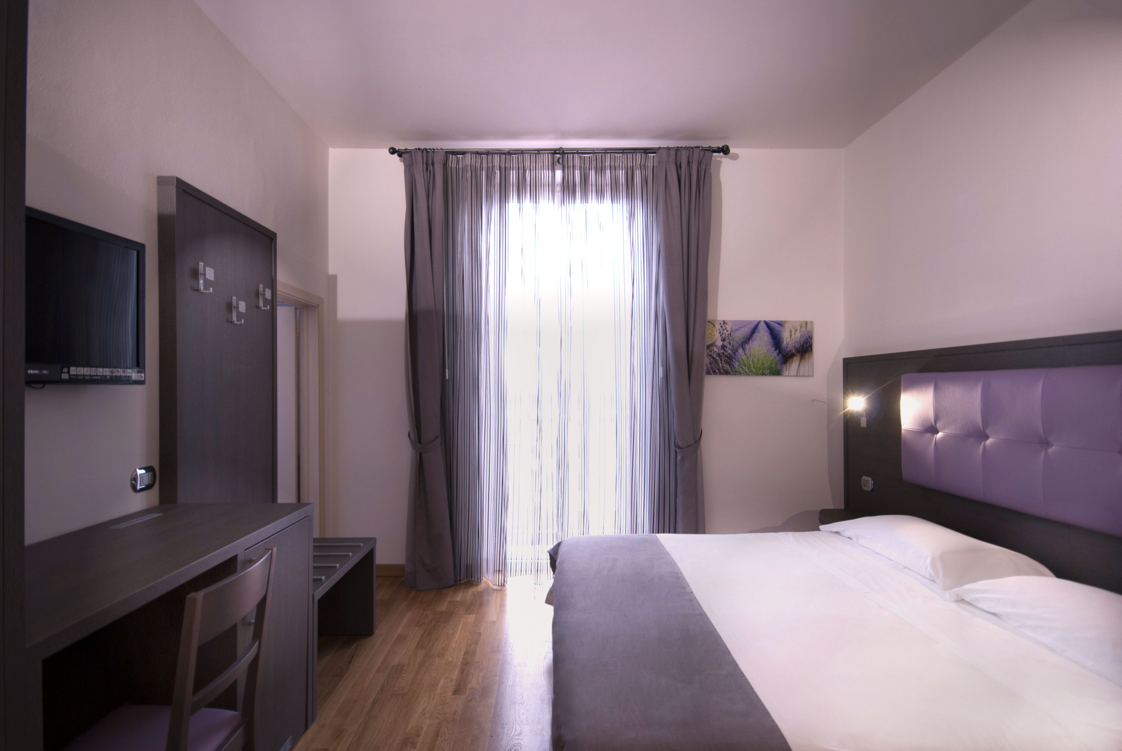 A hotel room with a bed, a desk, a TV, and a painting on the wall.