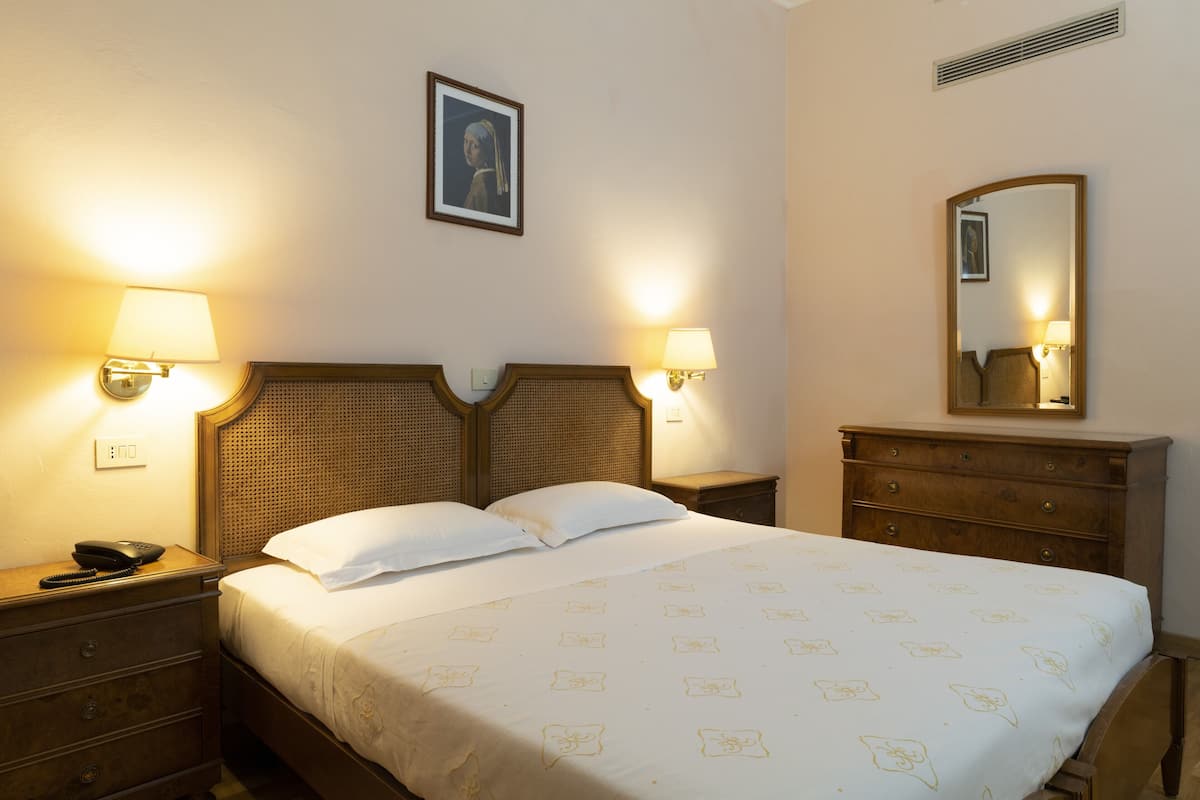 Une chambre d’hôtel comprenant un lit, deux tables de chevet, une petite table, une commode, un miroir et un tableau encadré au mur.