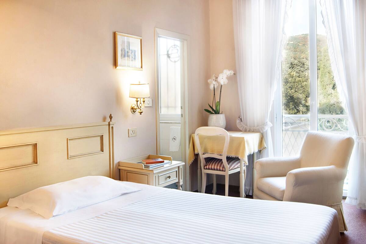Une chambre d’hôtel avec un lit, un bureau et une chaise, une fenêtre avec des rideaux et une vue sur la verdure.