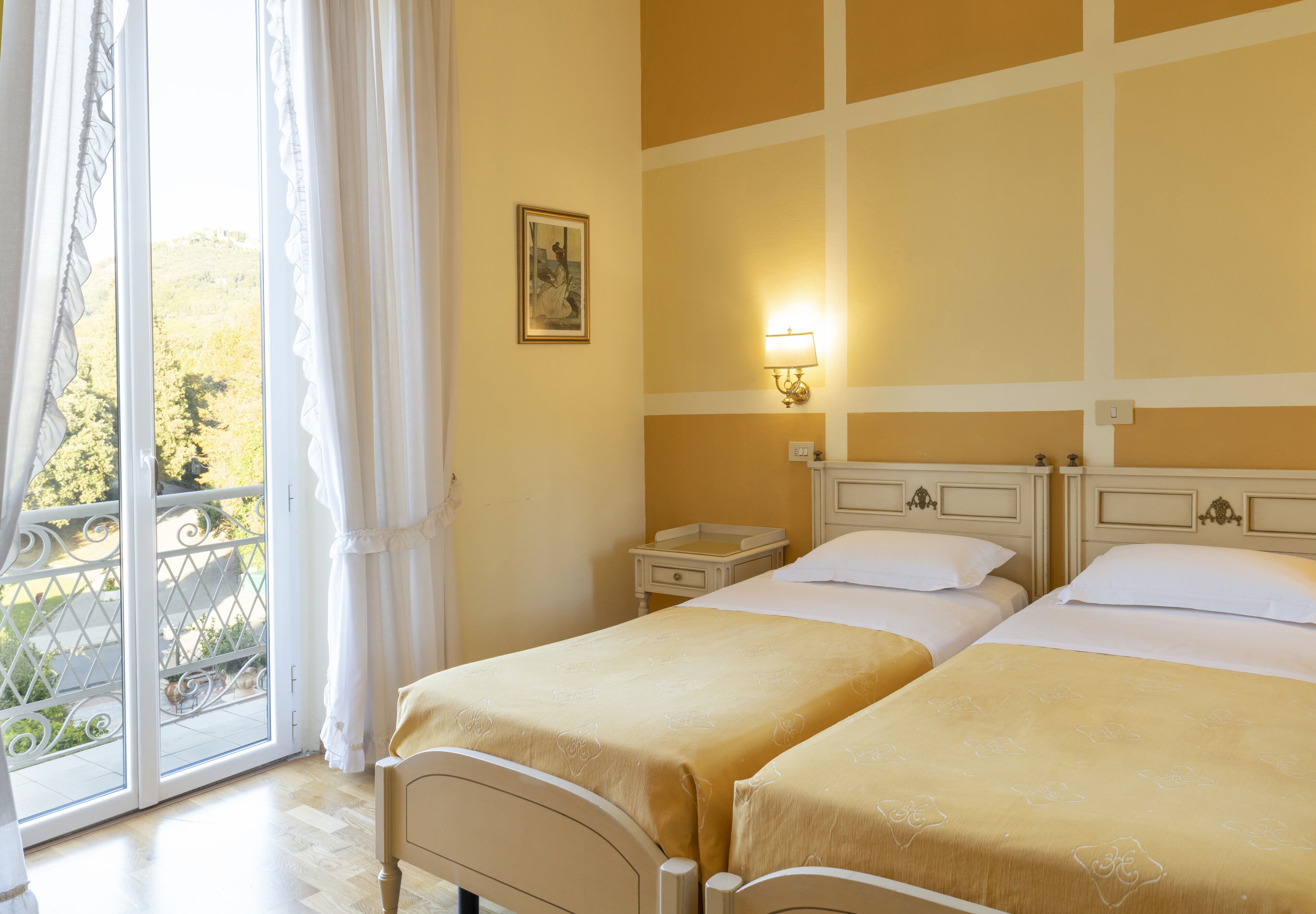 Une chambre d’hôtel avec deux lits, un balcon offrant une vue, un tableau accroché au mur et une table de chevet avec une lampe.