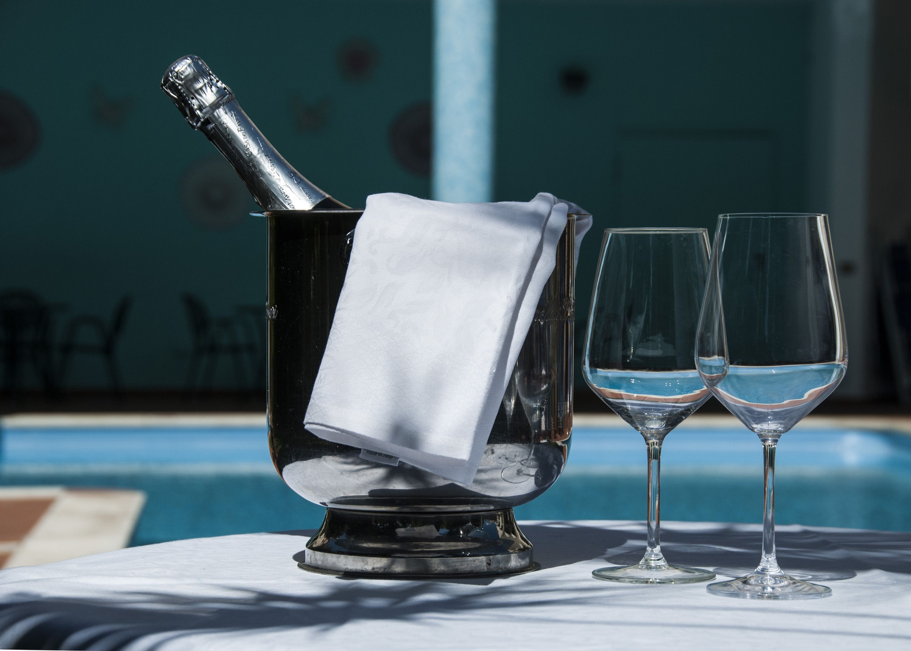 A champagne bottle in a metallic holder with a white napkin, two empty wine glasses, and a poolside table.