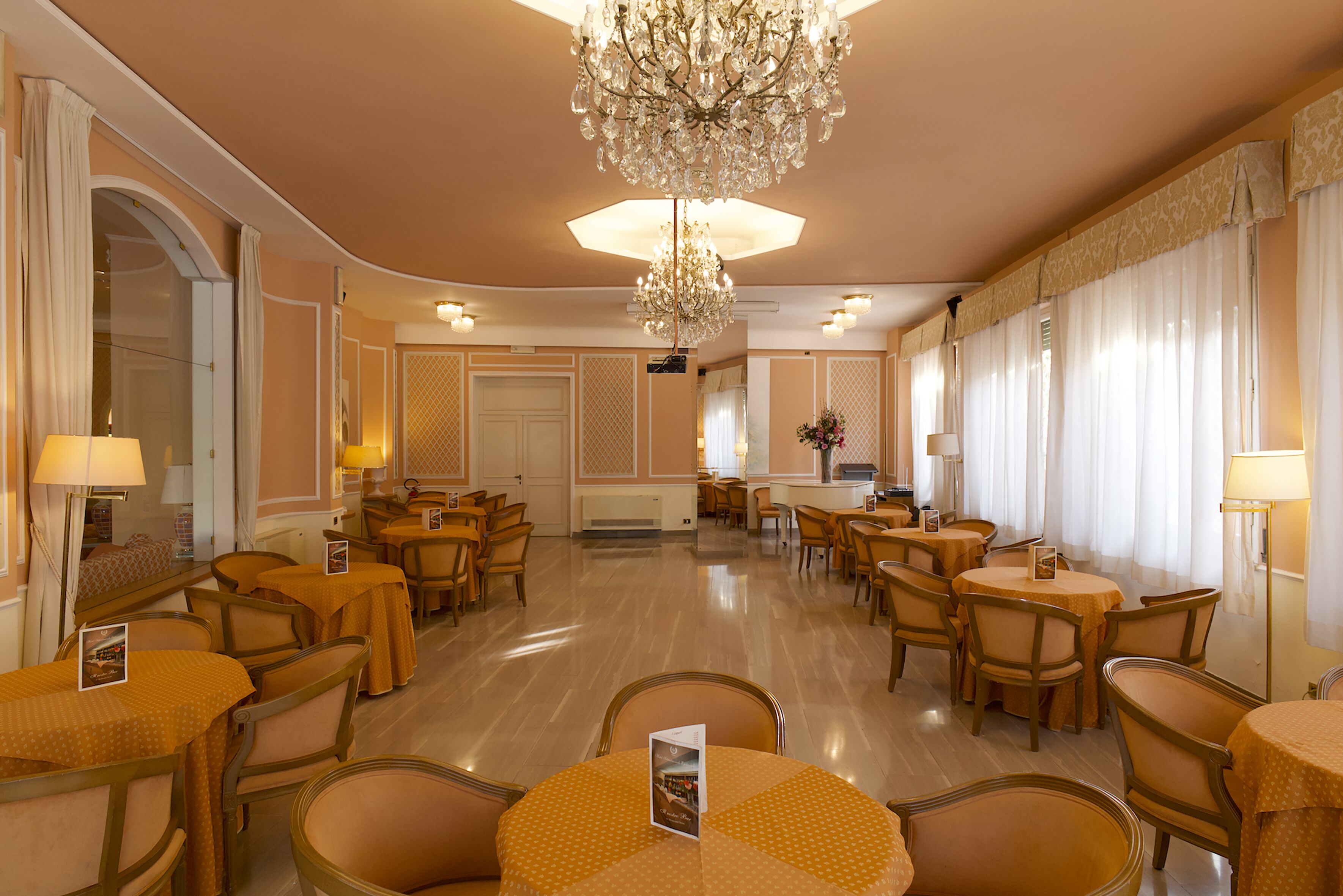 A spacious dining area with round tables and chairs, a chandelier, and a large mirror.