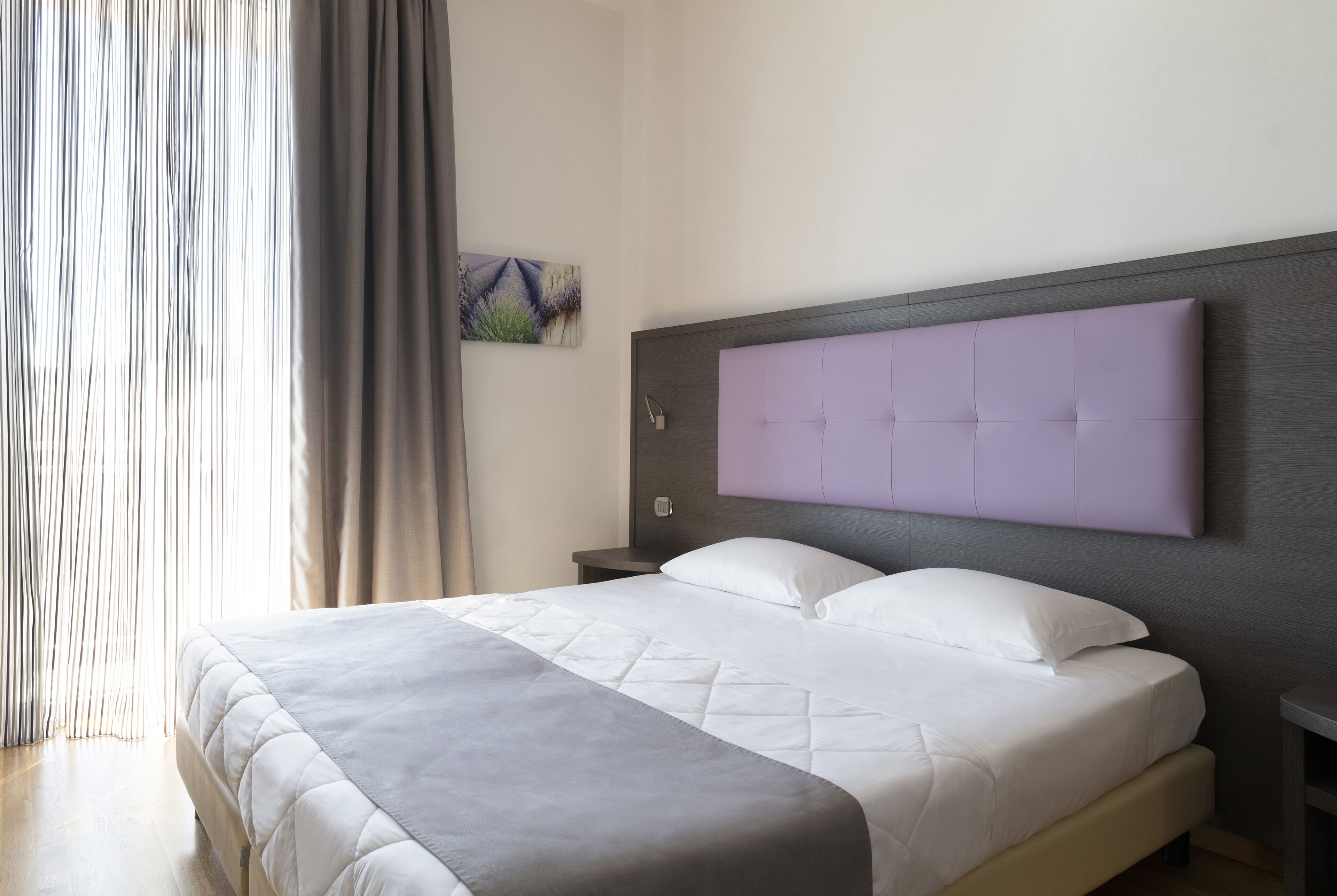 A neatly made bed with a grey and white quilt, a headboard with a purple panel, a nightstand with a lamp, and a painting on the wall.