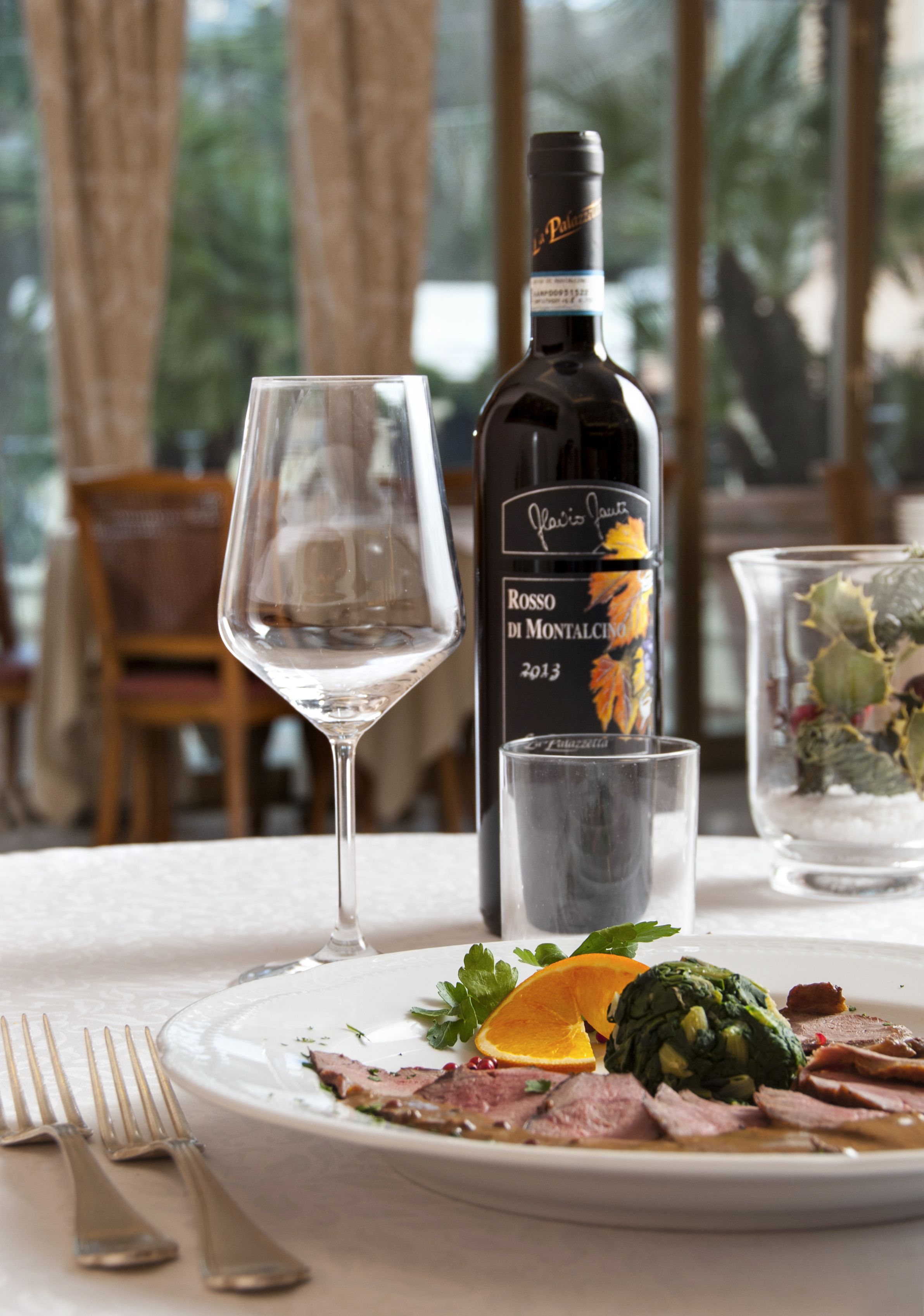 A dining table with a plate of food, a wine bottle, a wine glass, and a small glass with a flower arrangement.