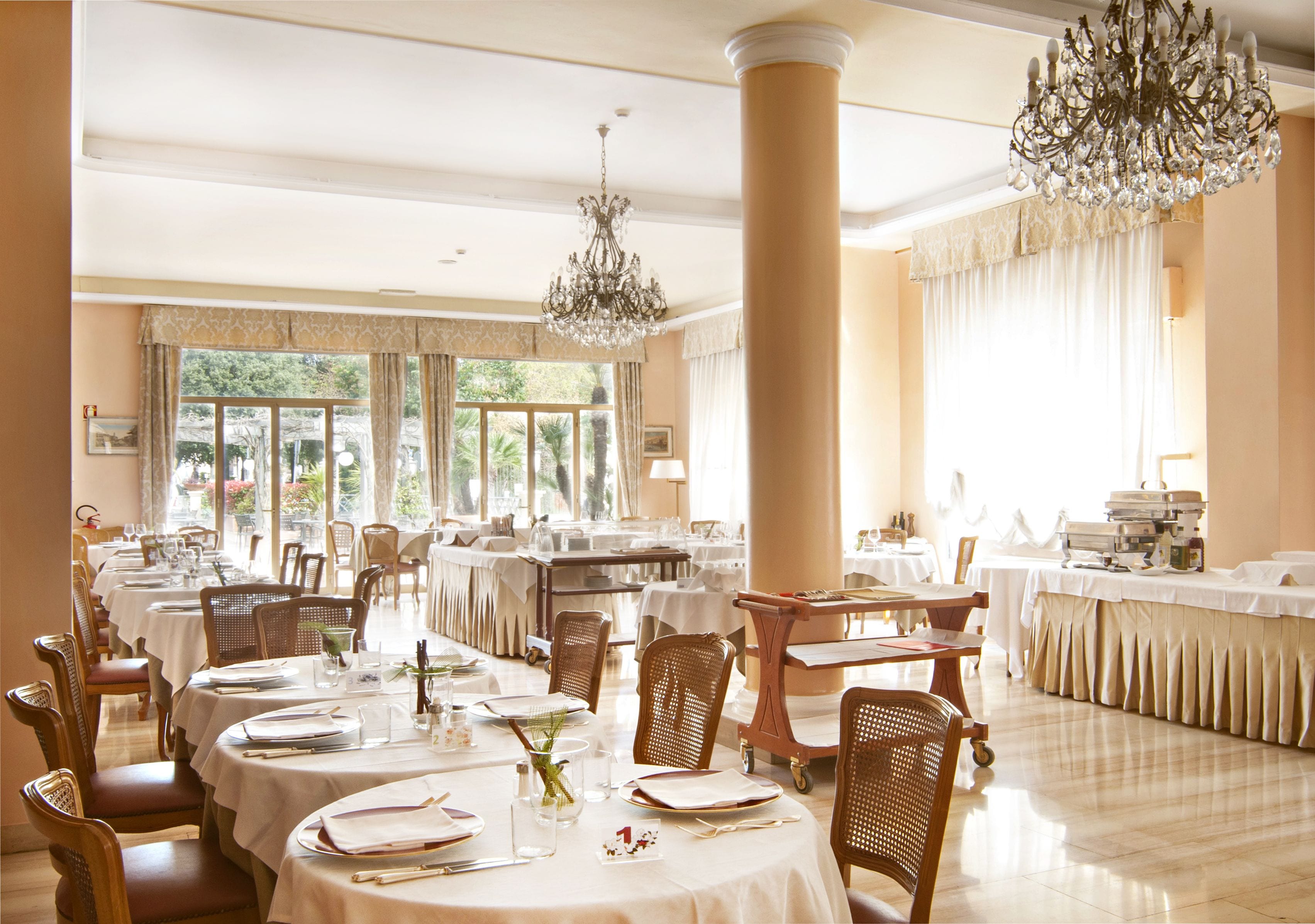 A dining area with round tables, chairs, chandeliers, and a large window with curtains.
