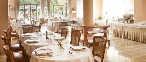 A dining area with round tables, chairs, chandeliers, and a large window with curtains.