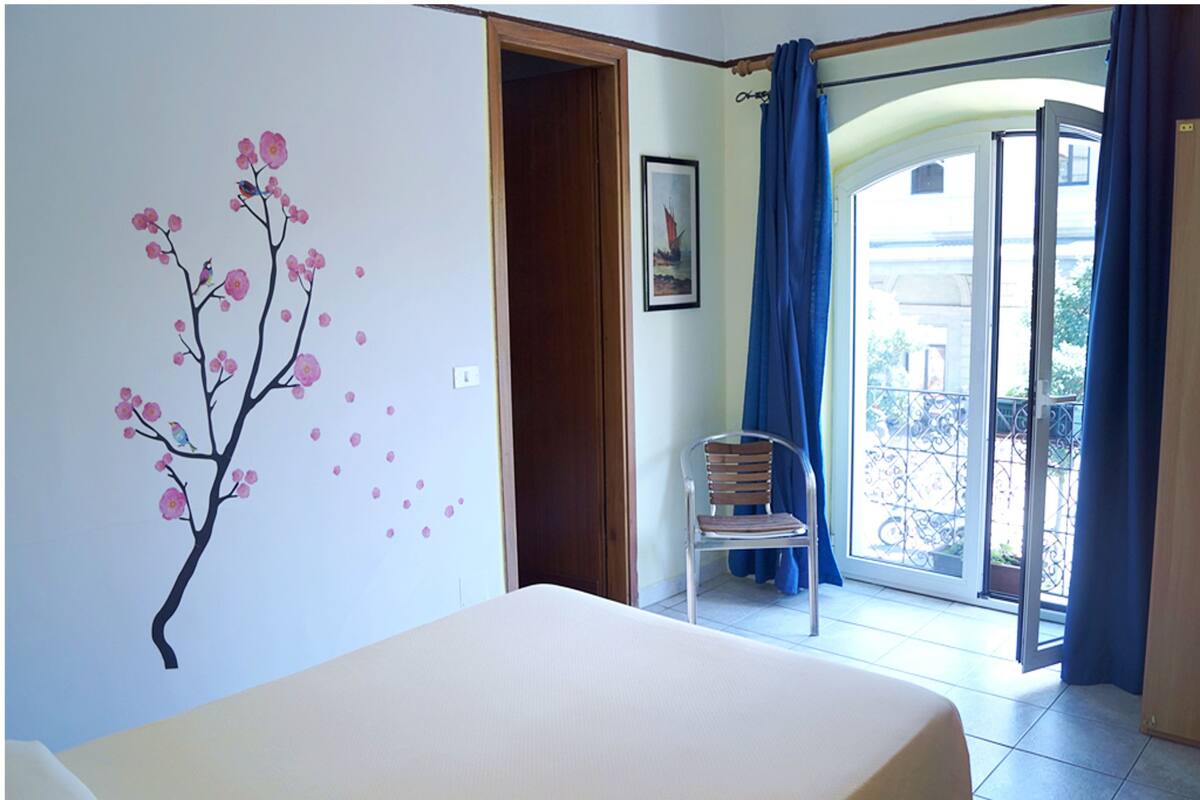 A room with a bed, a chair, a wall with a cherry blossom mural, a door, and a balcony with a railing.