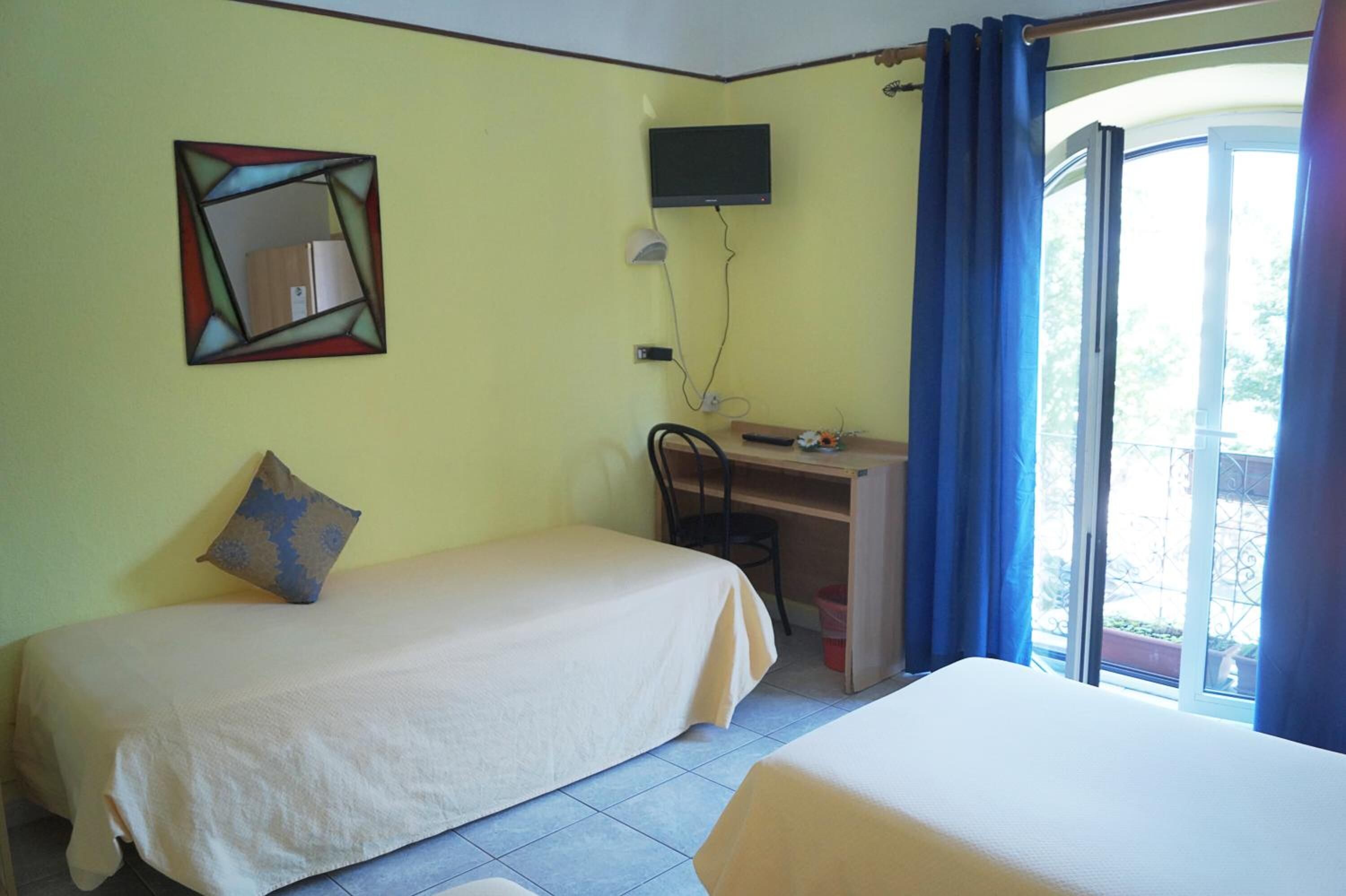 Ein Zimmer mit zwei Betten, einem Schreibtisch mit Computer, einem an der Wand montierten Fernseher und einem Fenster mit Vorhängen.