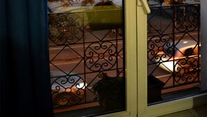 A view through a window with a metal railing, a building with multiple stories, and a tree with autumn leaves.