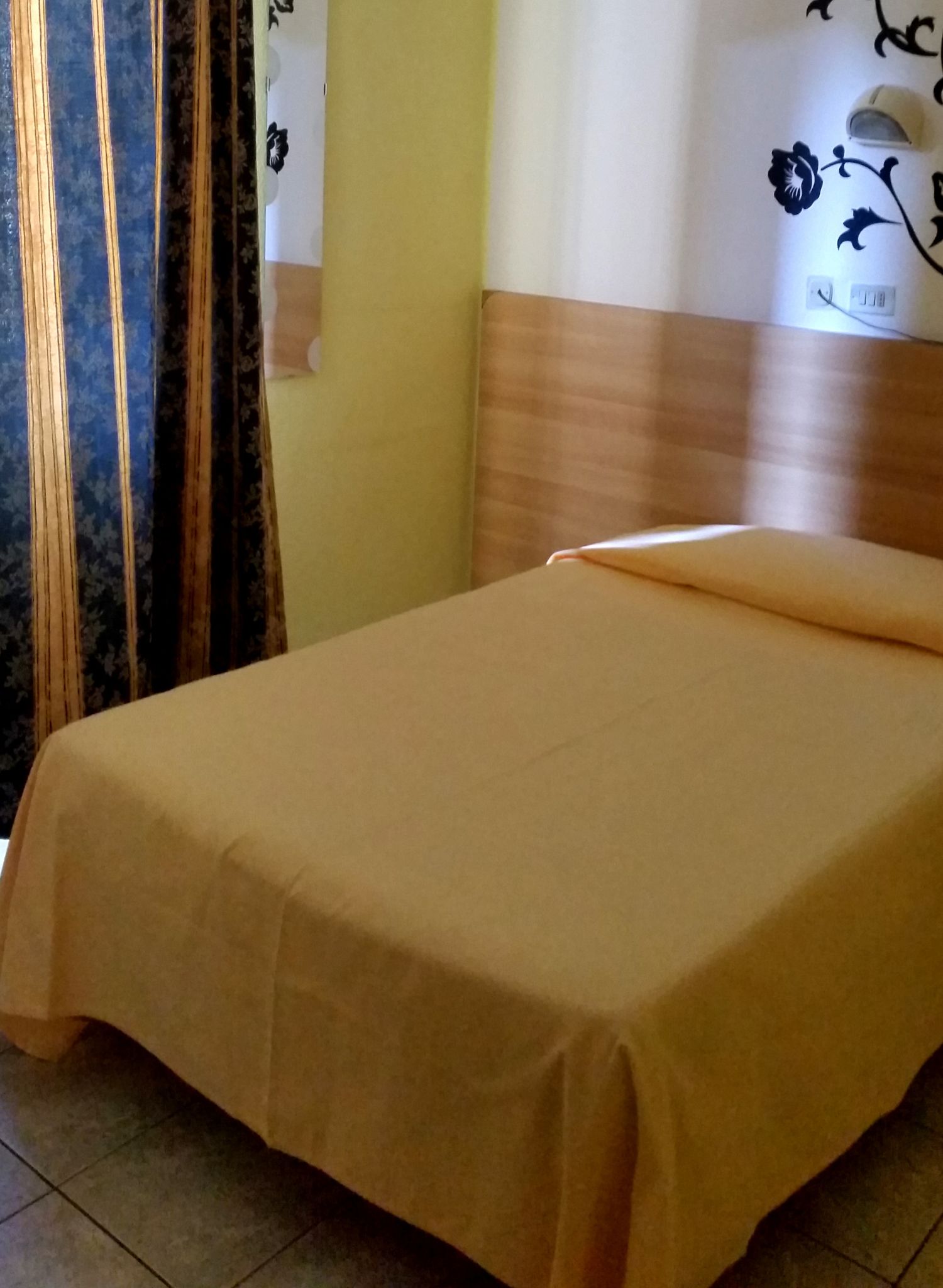 Ein ordentlich bezogenes Bett mit einer beigen Tagesdecke in einem Raum mit einem Holz-Kopfstück, einer Wand mit Blumenmuster und einem Vorhang mit floralem Design.
