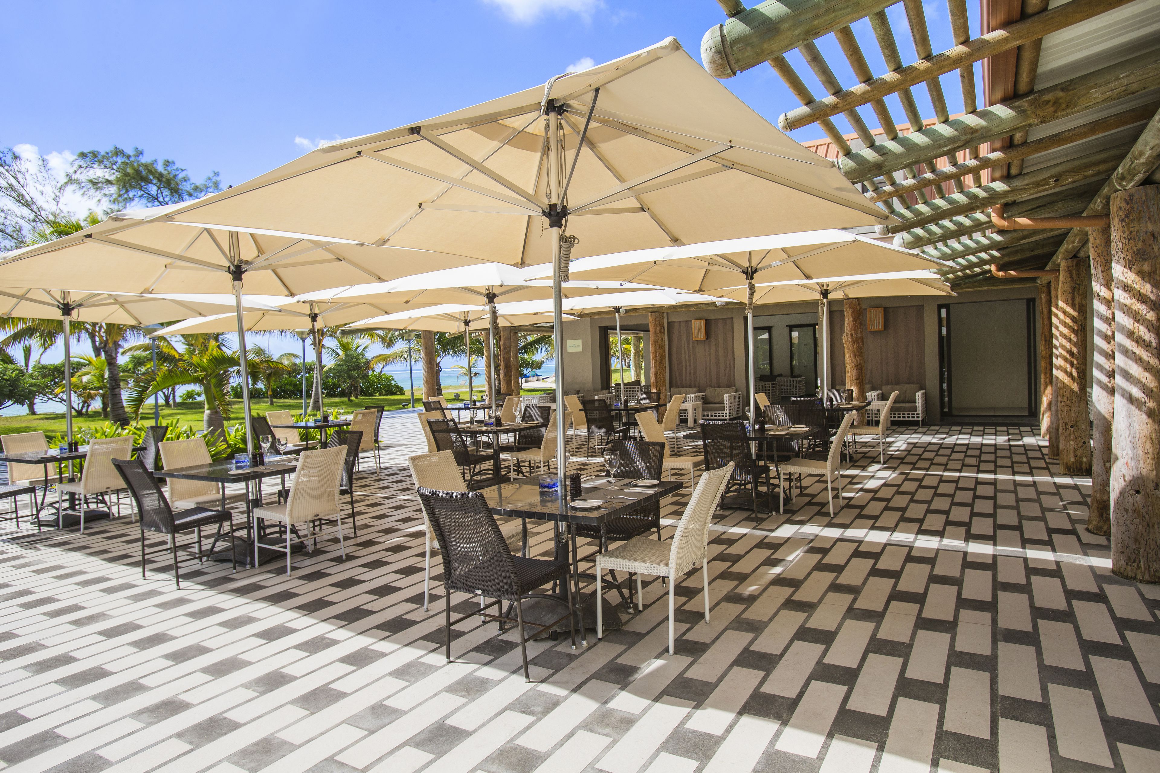 Un patio exterior con mesas y sillas bajo sombrillas grandes, suelo a cuadros y vistas al océano.